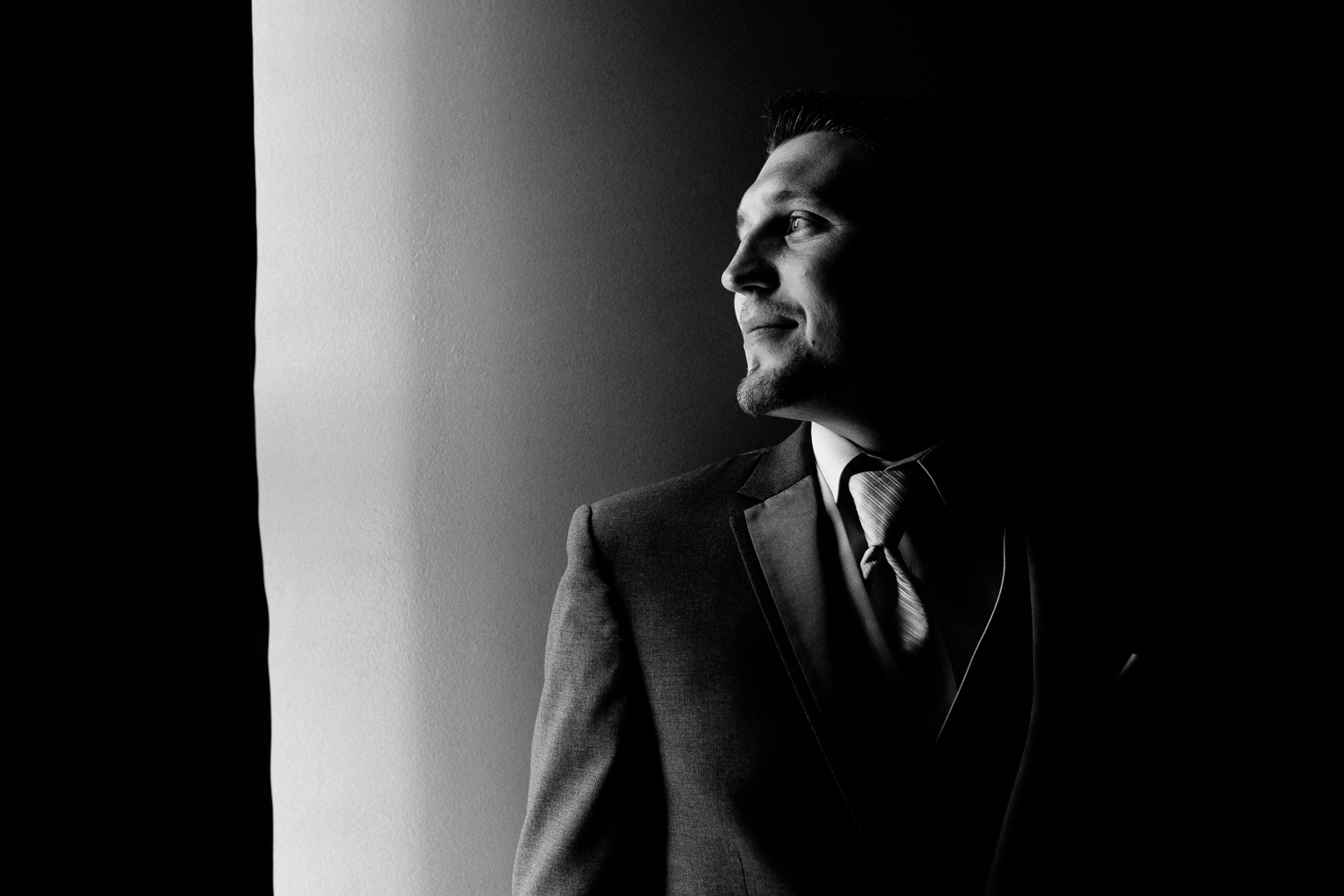  Black and white up close image of a groom looking out a window. He is smiling slightly. 