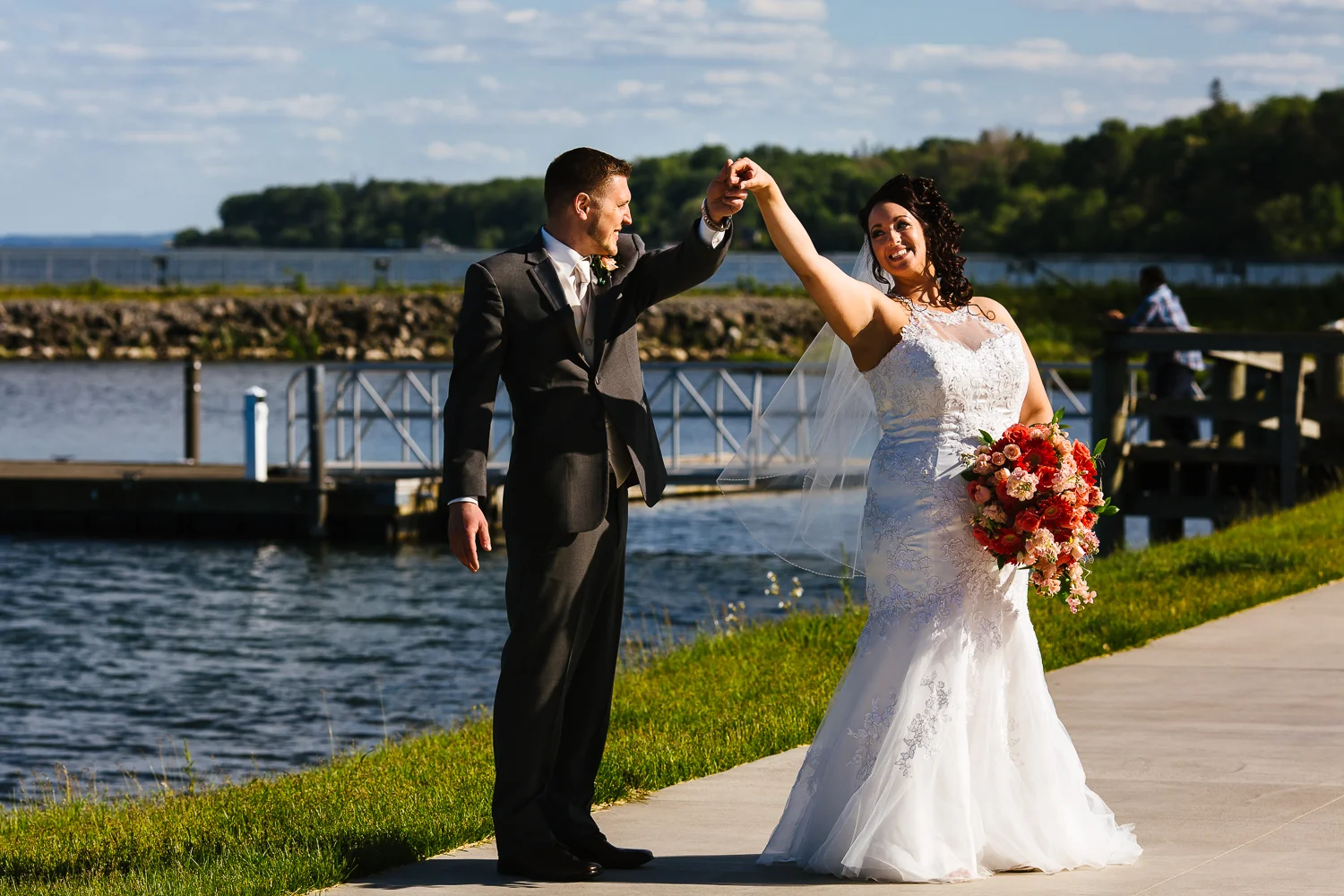 Bride looks back at groom outside near a lake. A man is fishing in the background.