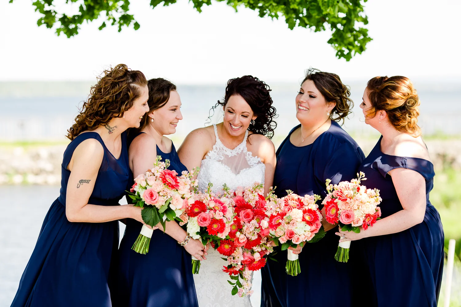 Bridesmaids wearing navy blue dresses surround a bride in a white gown. They have coral flowers. They are laughing.