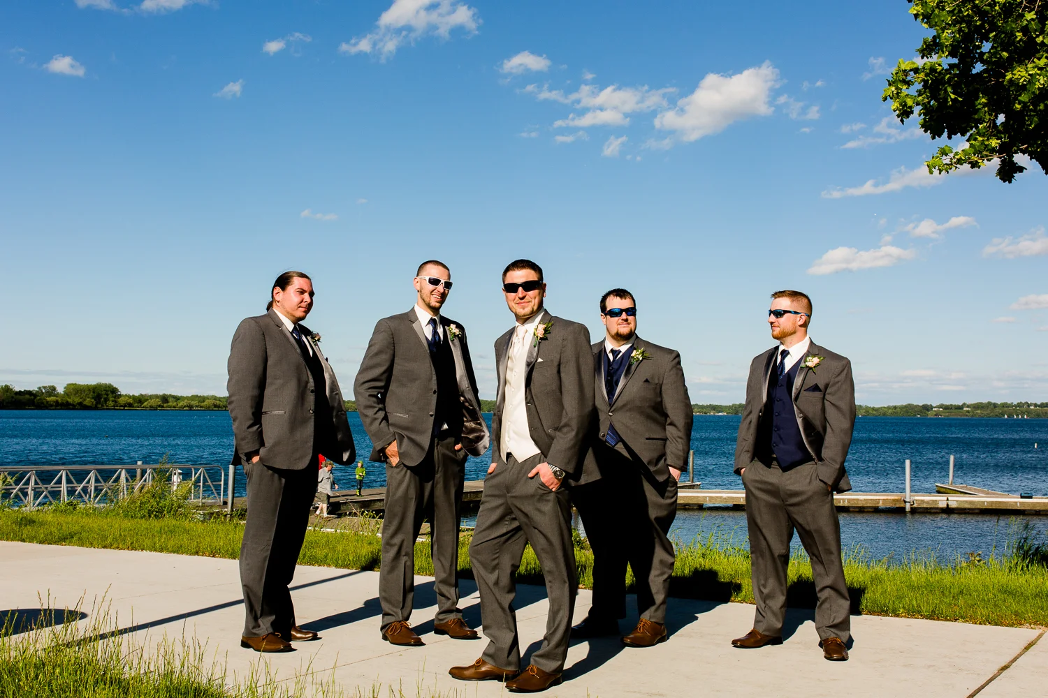 The groom and groomsmen have their backs to a lake. They are wearing sunglasses. There are clouds in the sky.