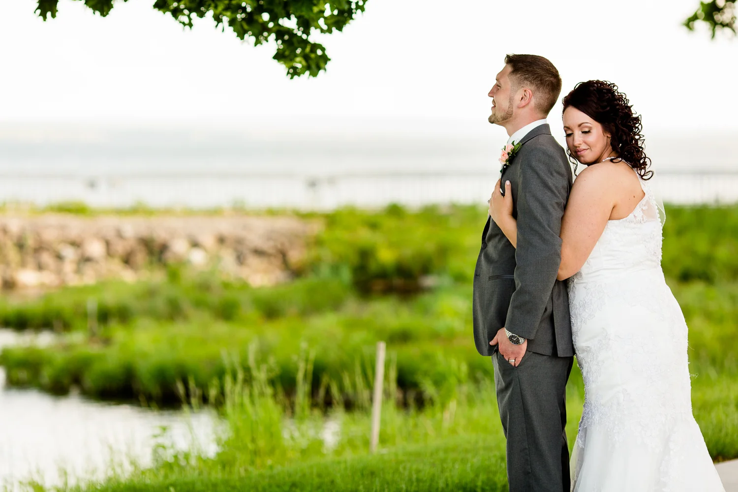 Bride holds groom from behind. Groom looks out into the distance. There is green grass behind them.