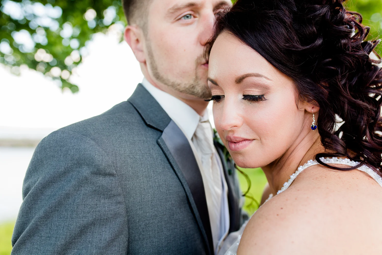 A close up of the bride's face. She has sapphire earrings on. The groom is kissing the side of her head.