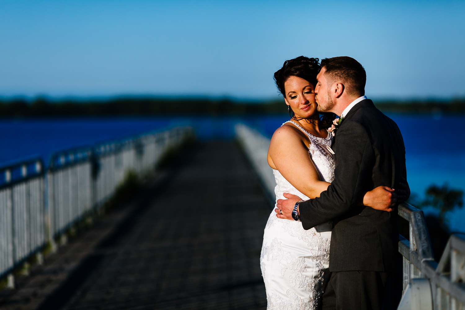  Groom kisses his bride on a pier at sunset. A blue lake is in the background. 