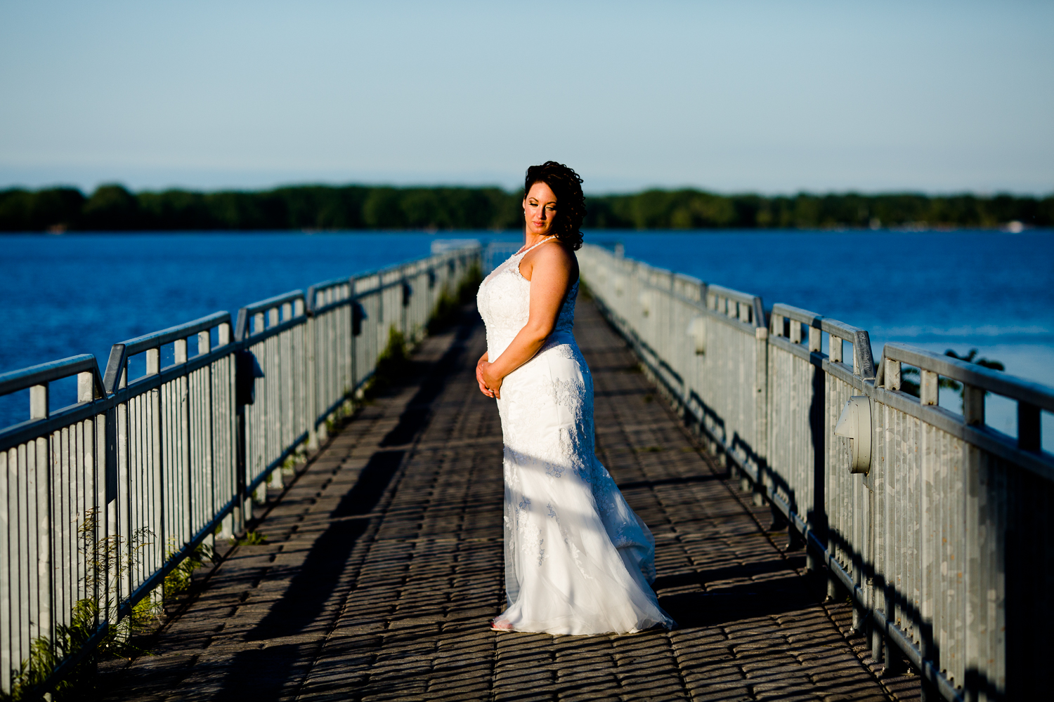  A bride in a white wedding dress stands in the center of a pier at sunset. The blue lake surrounds the pier. She looks over her shoulder. 