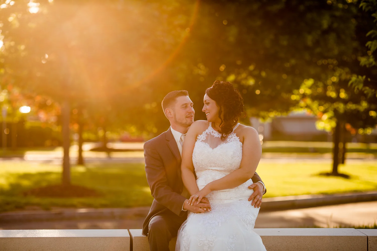 Bride and groom look at each other sweetly. There is sun flare in the image. The bride is sitting on the grooms lap.
