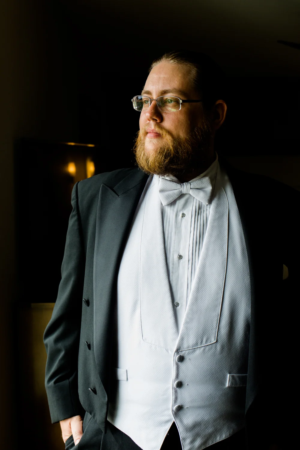 Portrait of a groom looking out a window. He has a black tux on with a white bowtie.
