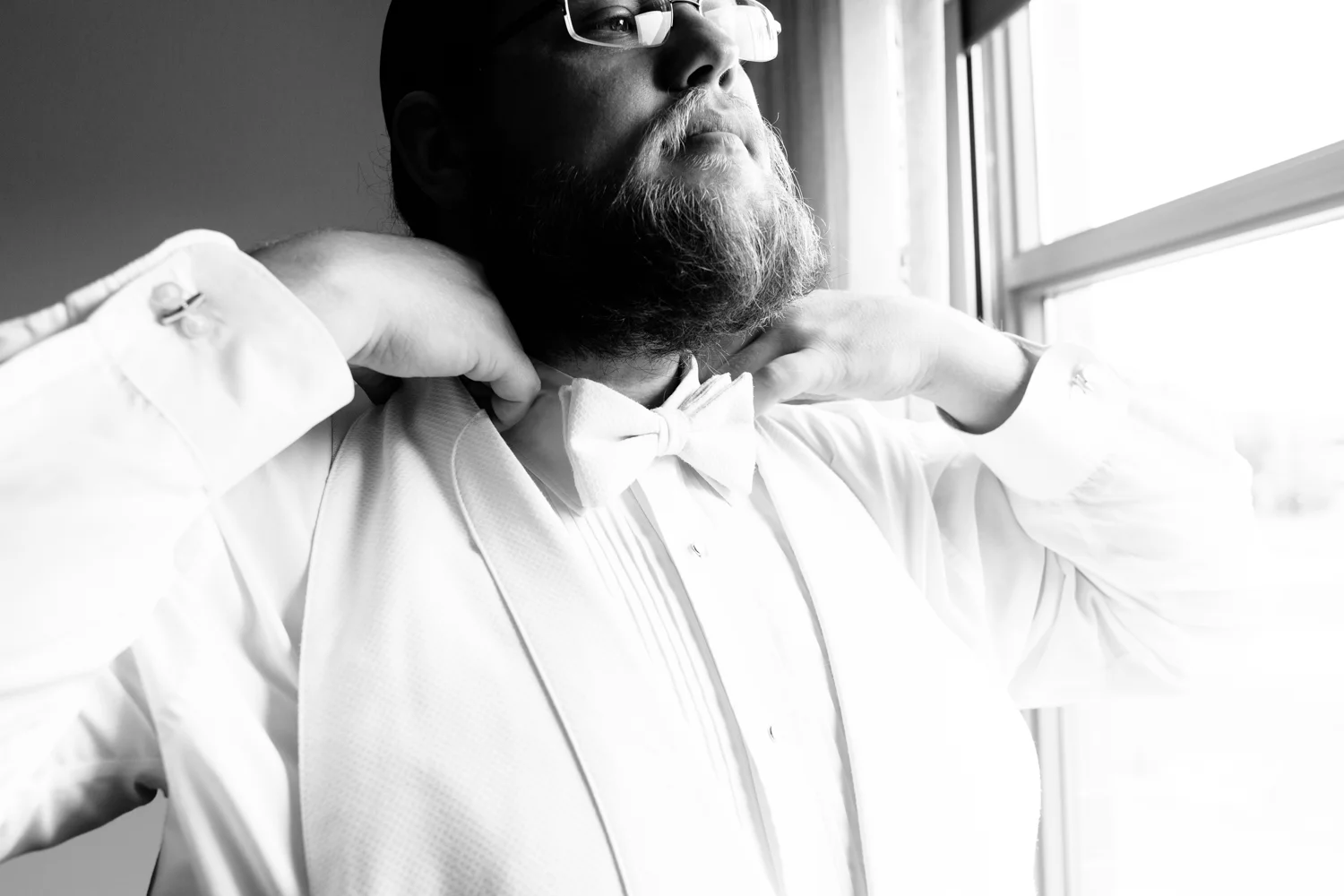 Black and white image of a groom adjusting his bowtie.