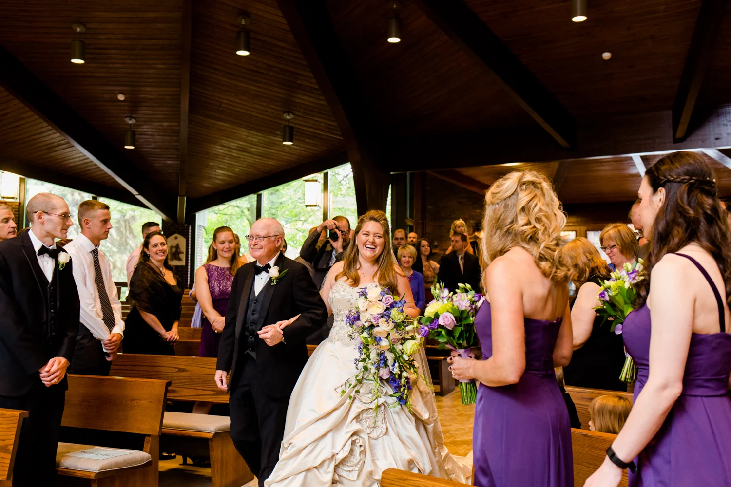 Bride walks down the aisle with her father. She is laughing and smiling at her bridesmaids. She is in a church.