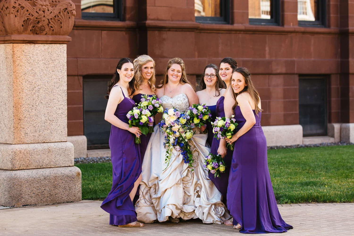 Bride in an Ivory wedding gown poses with bridesmaids in purple floor length dresses.