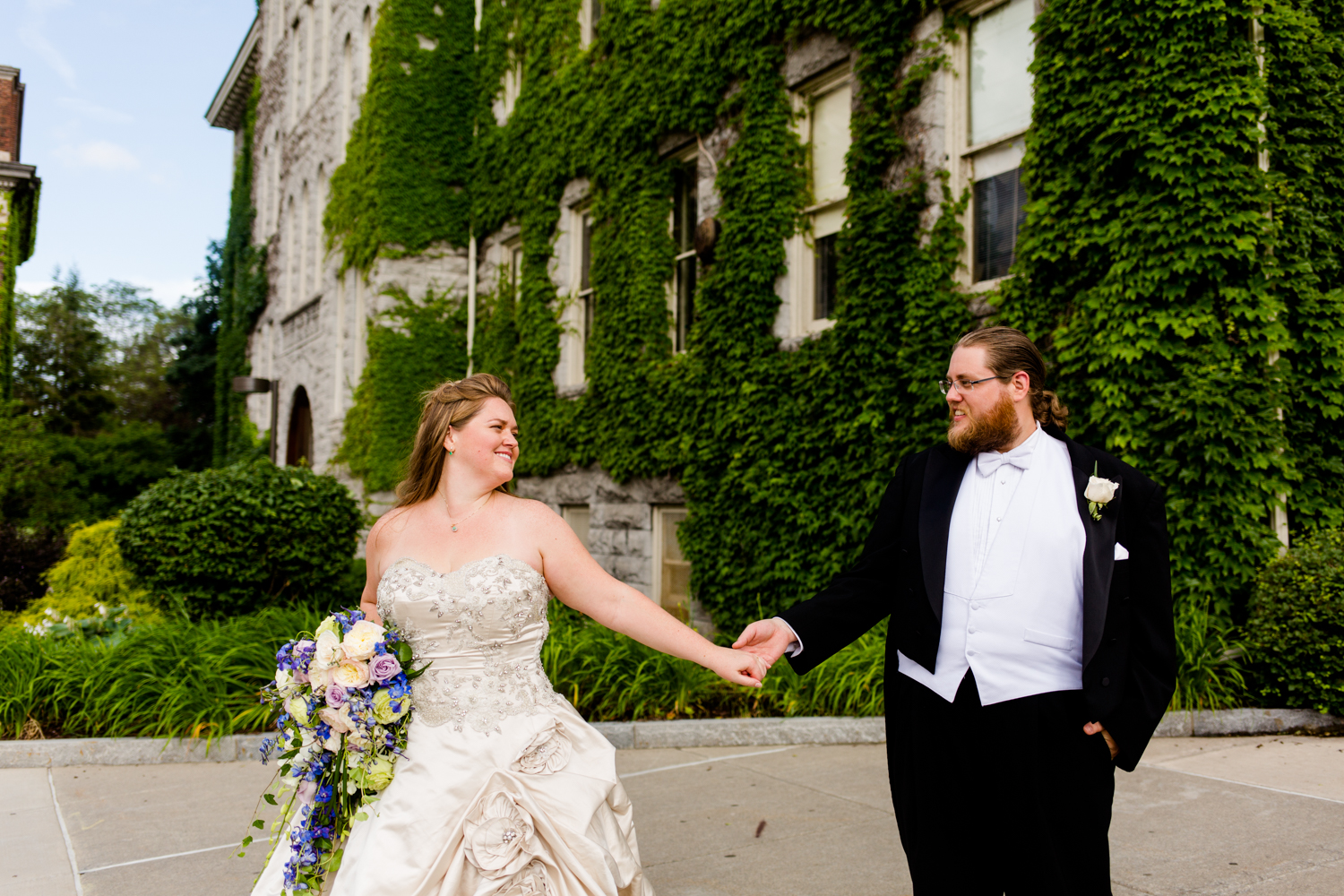 Portrait of the bride and groom. They are standing in front of a stone building with ivy growing on it. It is located at Syracuse University in Syracuse, NY.