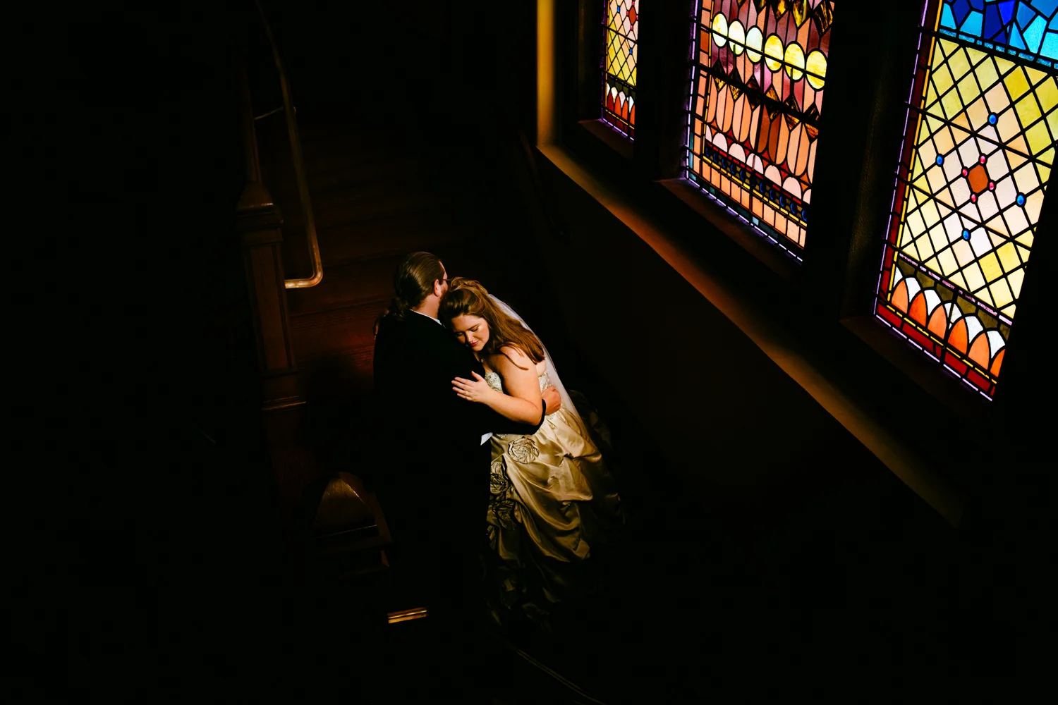 Moody portrait of bride leaning against groom. The background is dark except for a stainglassed window.