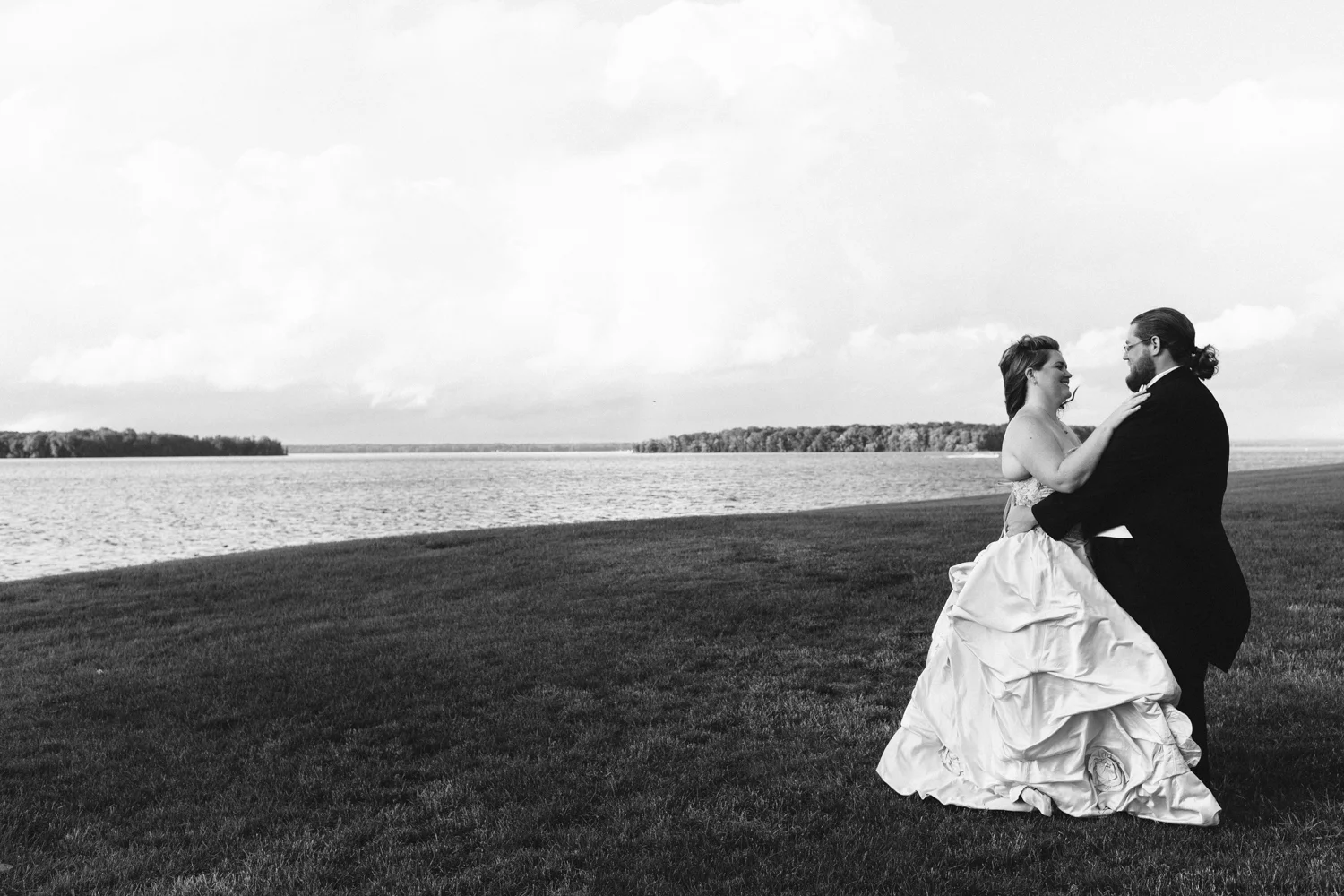Black and white image of a bride and groom standing near a lake. Bride is wearing a ball gown.