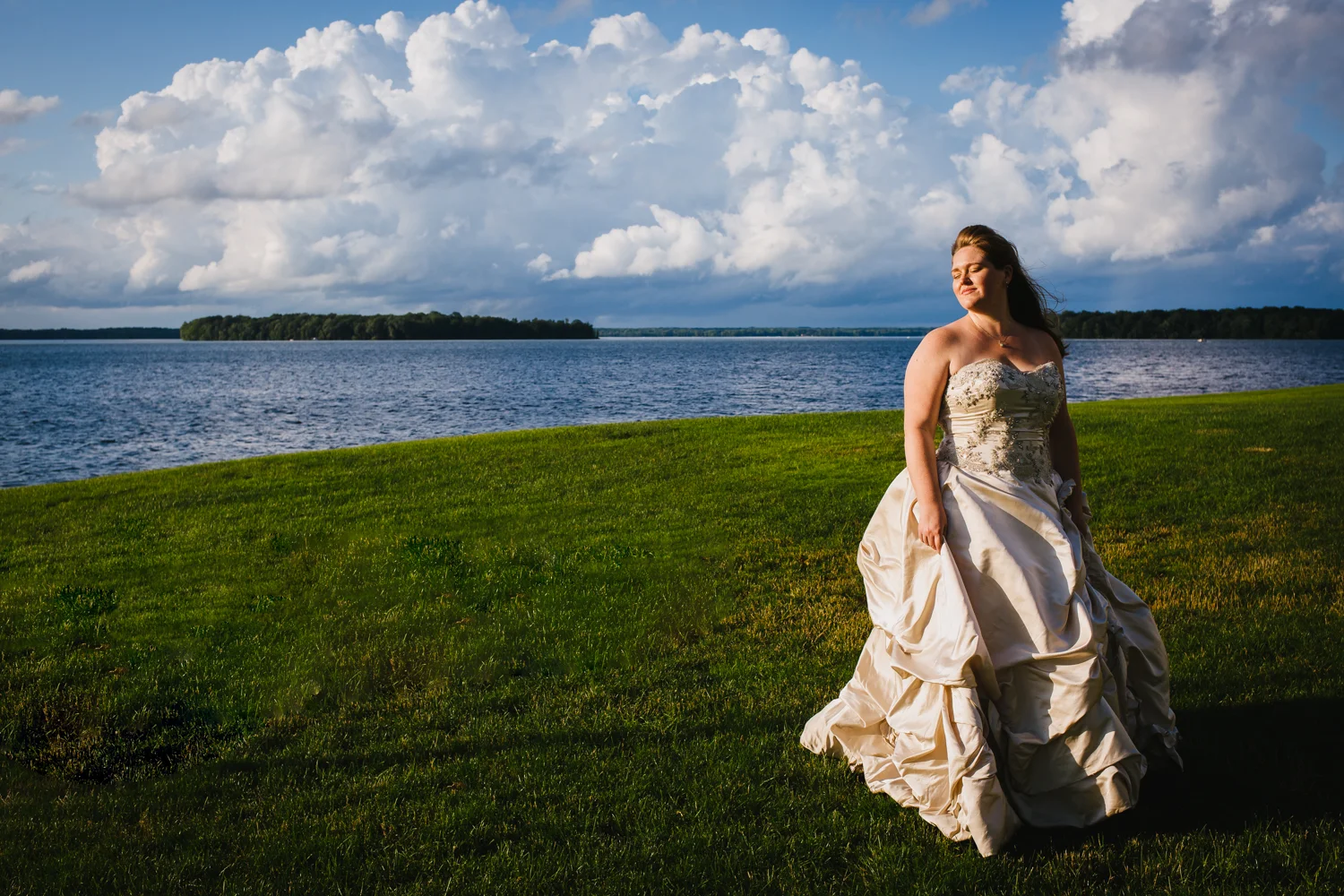 Bride smiles with eyes closed. She stands in front of a lake with fluffy white clouds behind her.