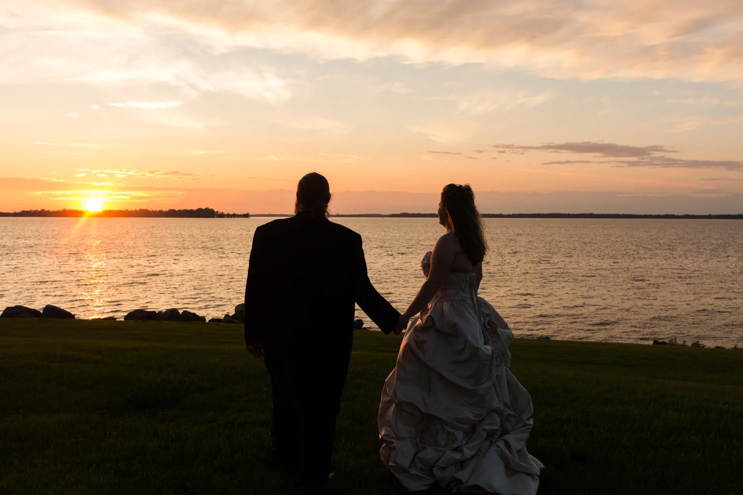 Bride and groom hold hands and watch the sunset across the lake.