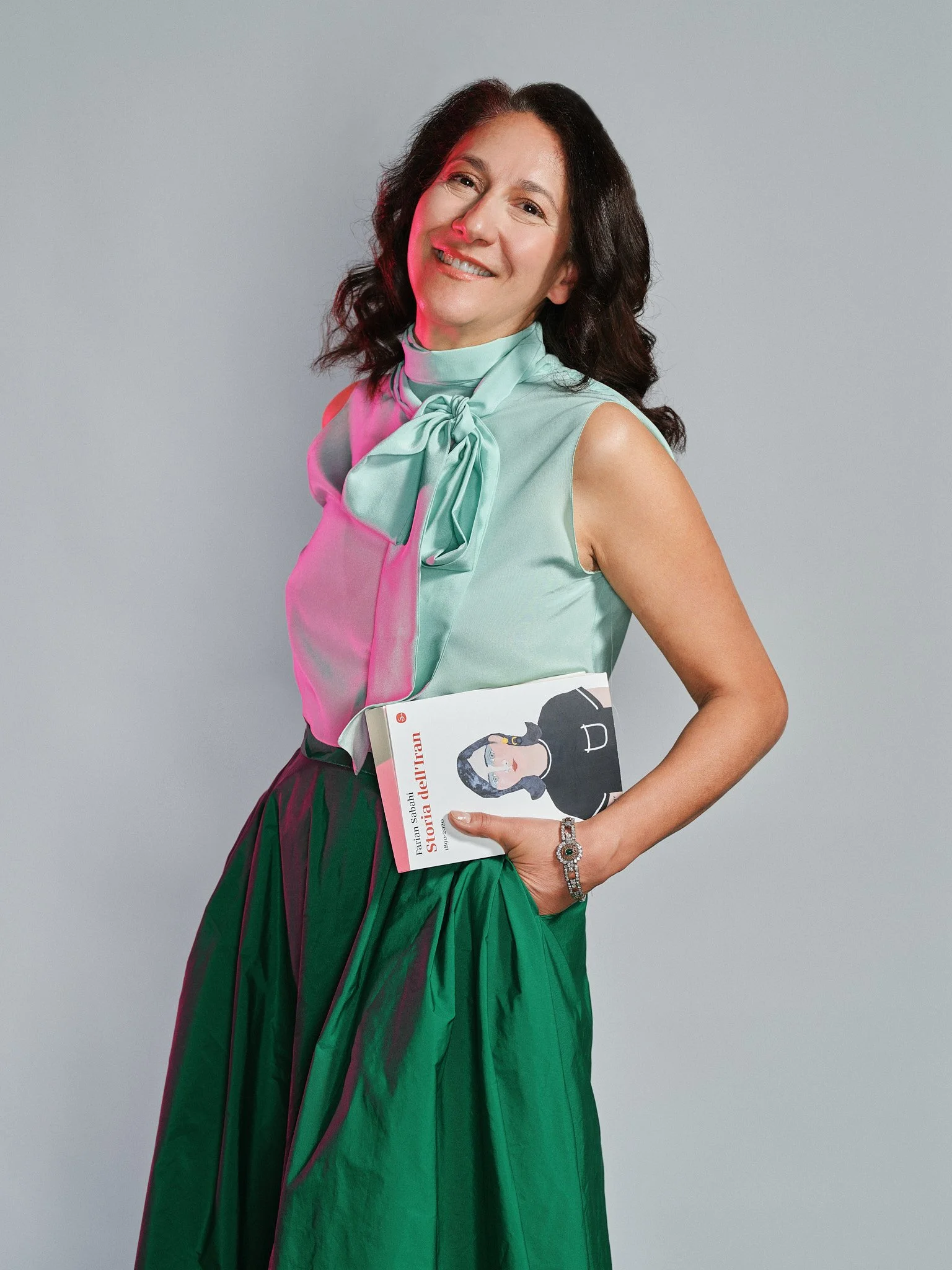 Donna con capelli castani ricci e sorriso, indossa una maglietta verde acqua con fiocco, una gonna lunga verde, e tiene un libro con illustrazione di una donna, sfondo grigio.