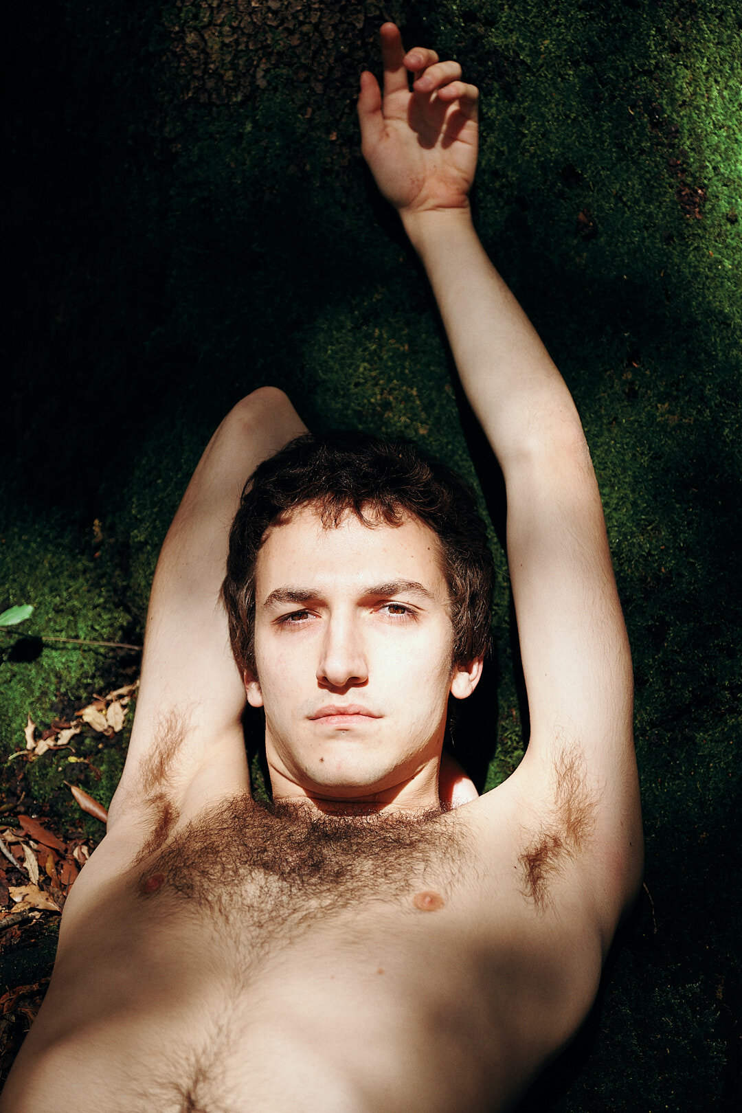 Ragazzo nudo sdraiato su muschio verde con un braccio sopra la testa e uno a fianco, guardando verso l'obiettivo in un ambiente naturale