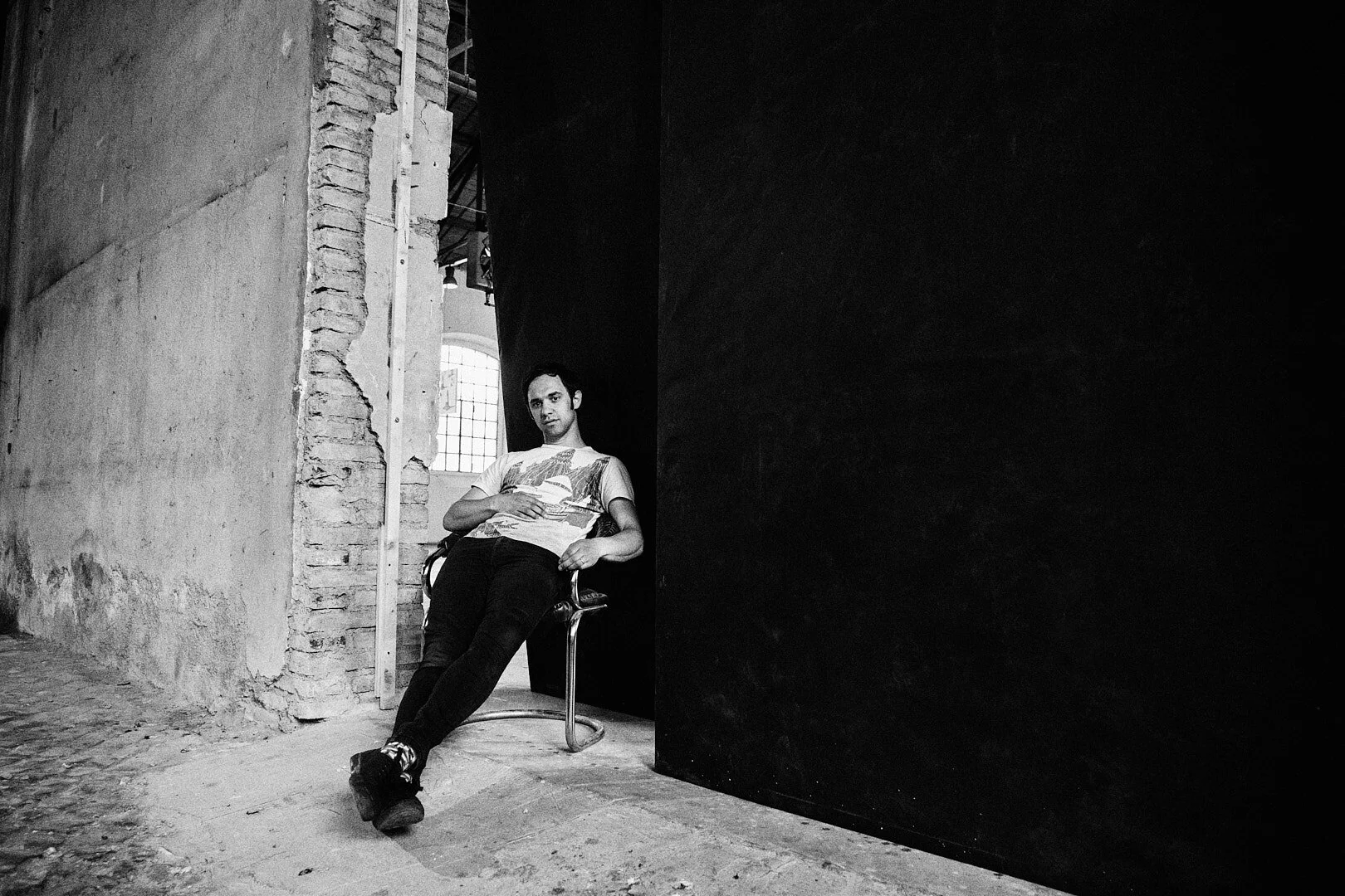 Ritratto del cantante Cosmo
Ragazzo seduto su una sedia, con pareti vuote e finestra in background.