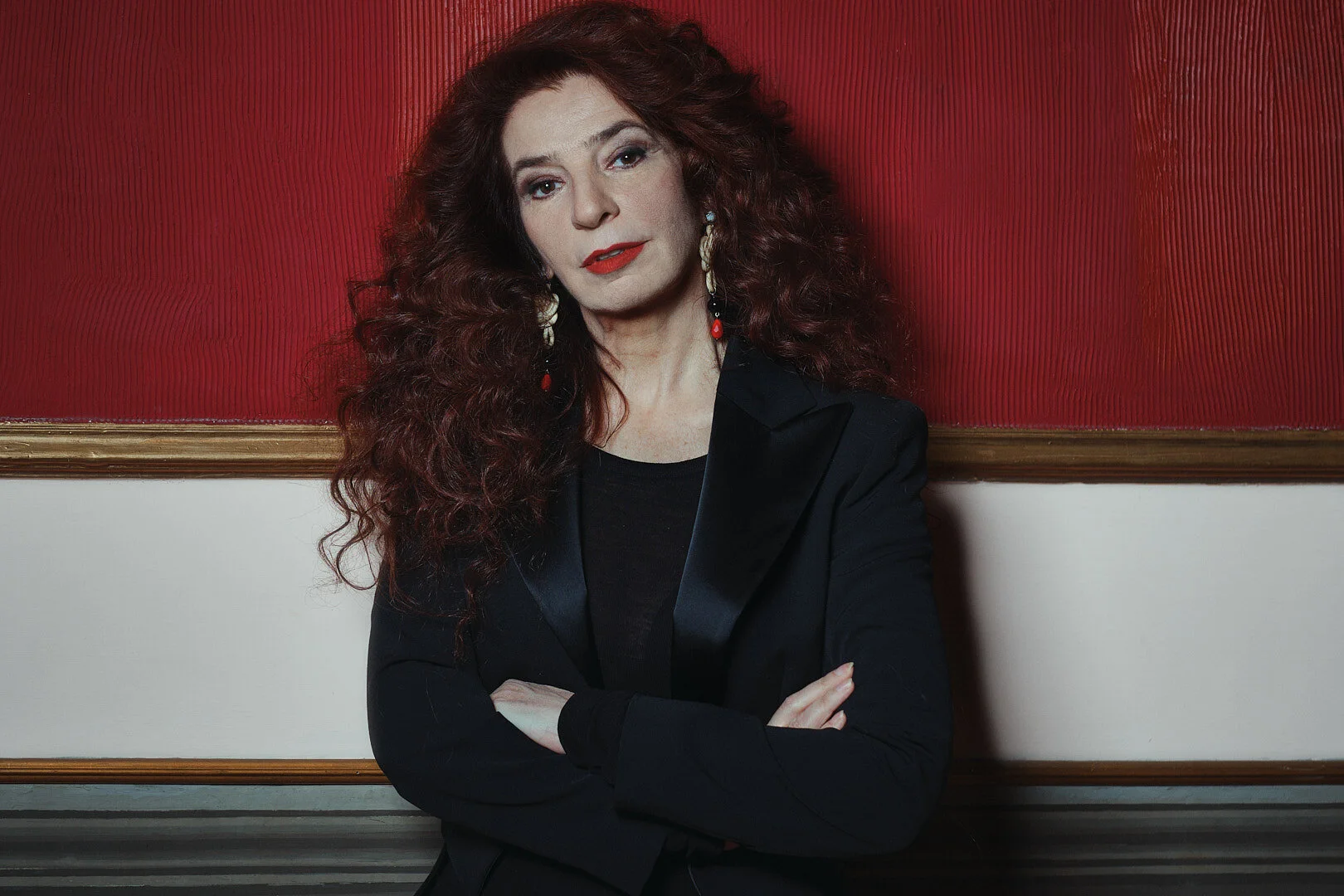 Attrice e Cantante Teresa De Sio
Una donna con capelli ricci di colore rosso scuro, indossa un completo nero, posa con le braccia incrociate di fronte a un muro rosso con decorazioni dorate.