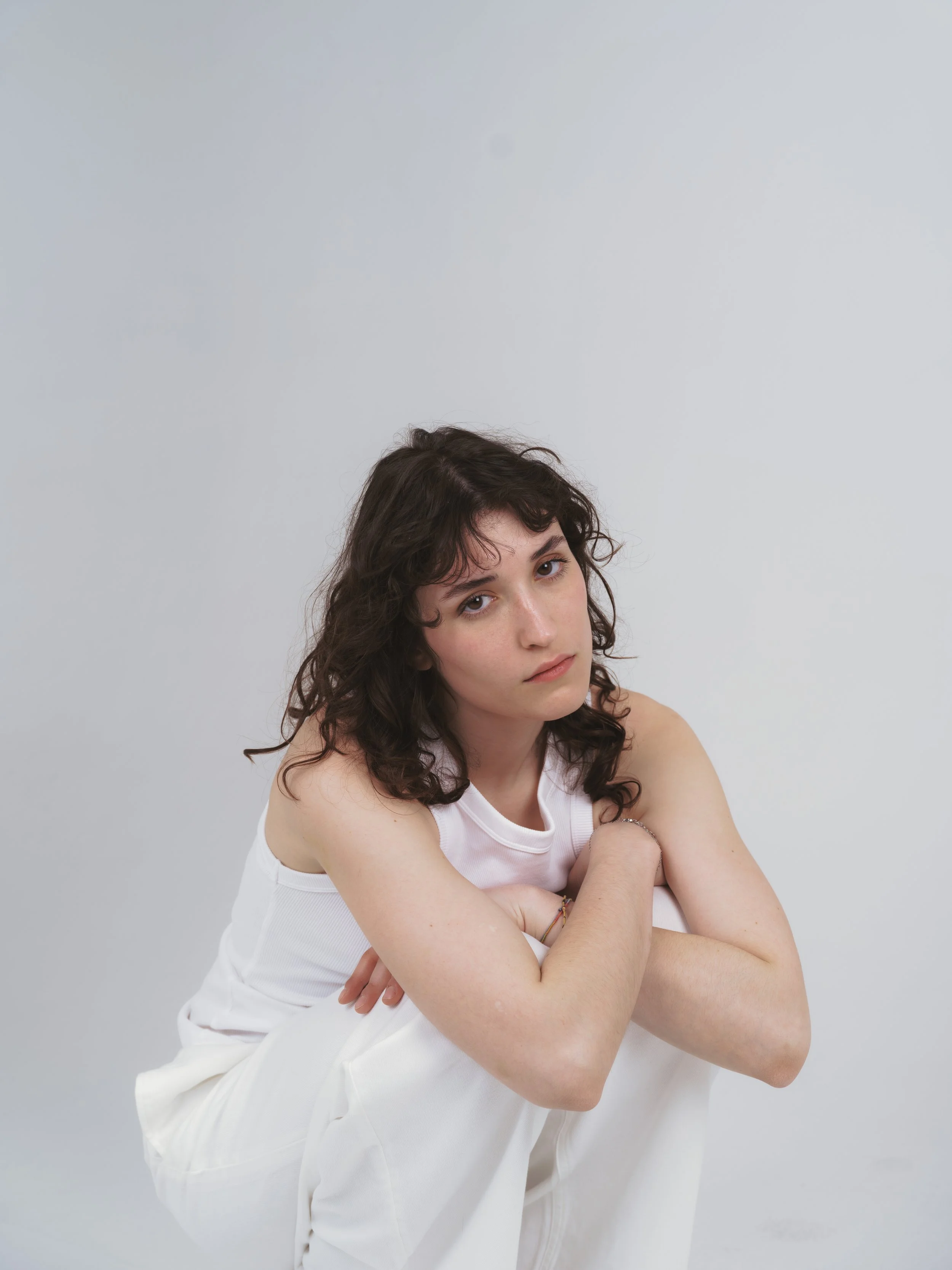 Ragazza con capelli castani ricci, vestita di bianco, con le braccia incrociate, che guarda verso destra su uno sfondo grigio chiaro.