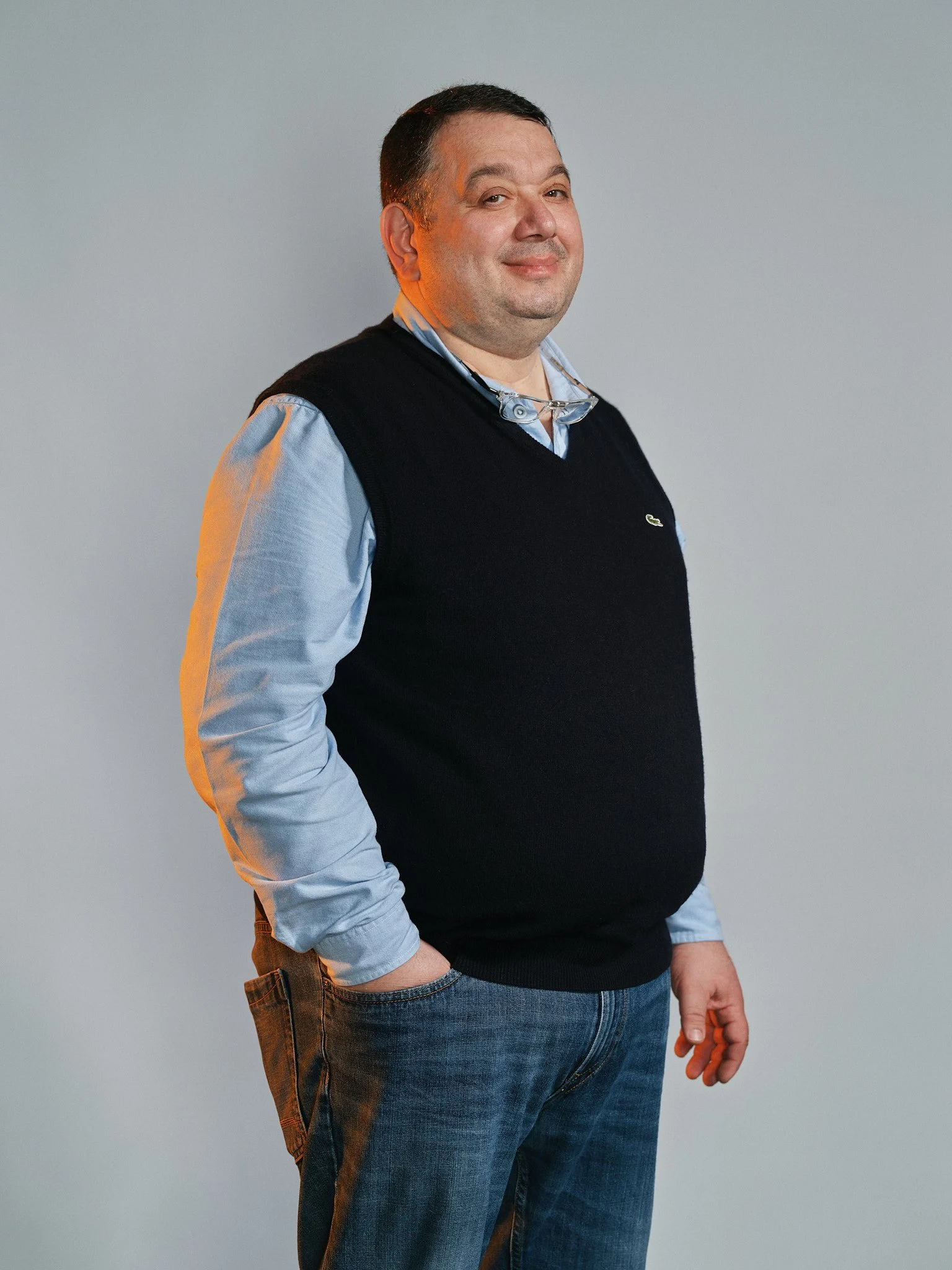 Uomo con maglione nero, camicia azzurra e jeans, che sorride leggermente, tiene degli occhiali da vista attorno al collo, su sfondo grigio.
