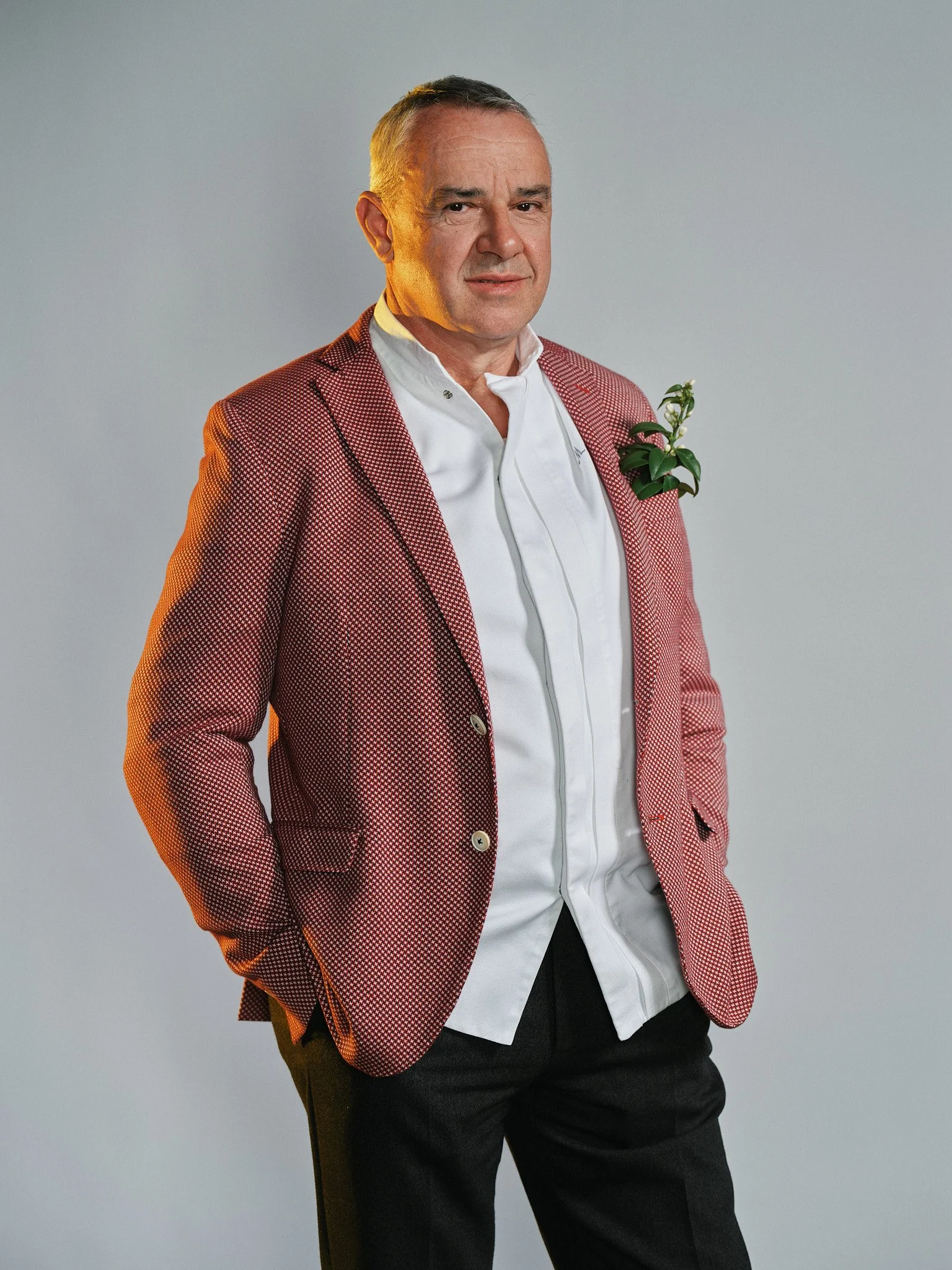 Uomo in abito elegante con fiore sulla giacca, posa con una mano in tasca, sfondo grigio.
