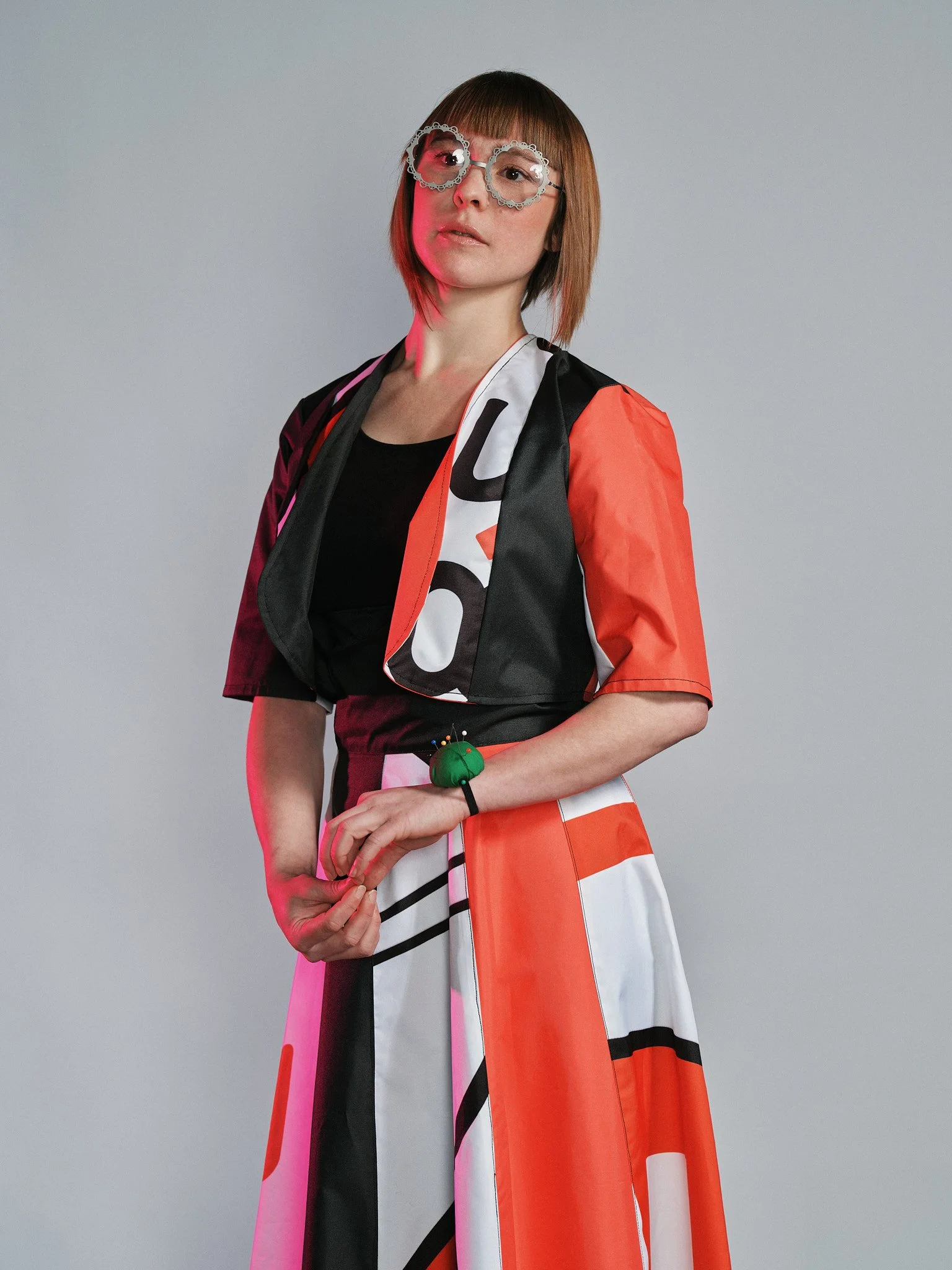 Una donna con capelli corti castano chiaro, indossa occhiali dal frame decorativo e un abito bianco, nero e rosso con il logo McDonald's. Tiene le mani incrociate davanti e sul polso porta un braccialetto verde con una toppa bianca. Sfondo grigio.