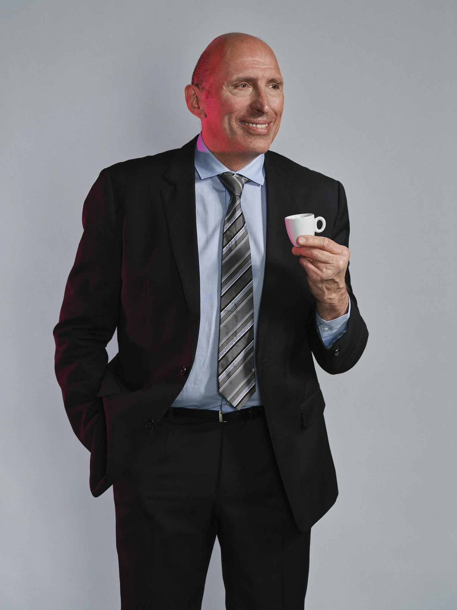 Uomo d'affari con abito nero, camicia azzurra e cravatta in piedi che tiene una tazza da caffè. Sfondo grigio.