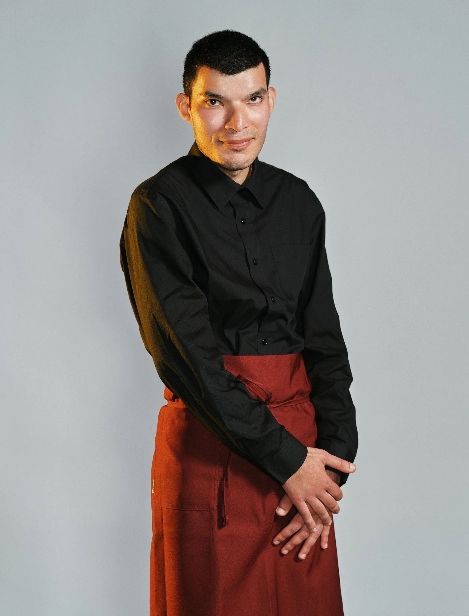 Ritratto di un uomo con i capelli corti, indossa una camicia nera e pantaloni di colore arancione, posa sulla fronte una mano con le dita intrecciate, sfondo grigio chiaro.