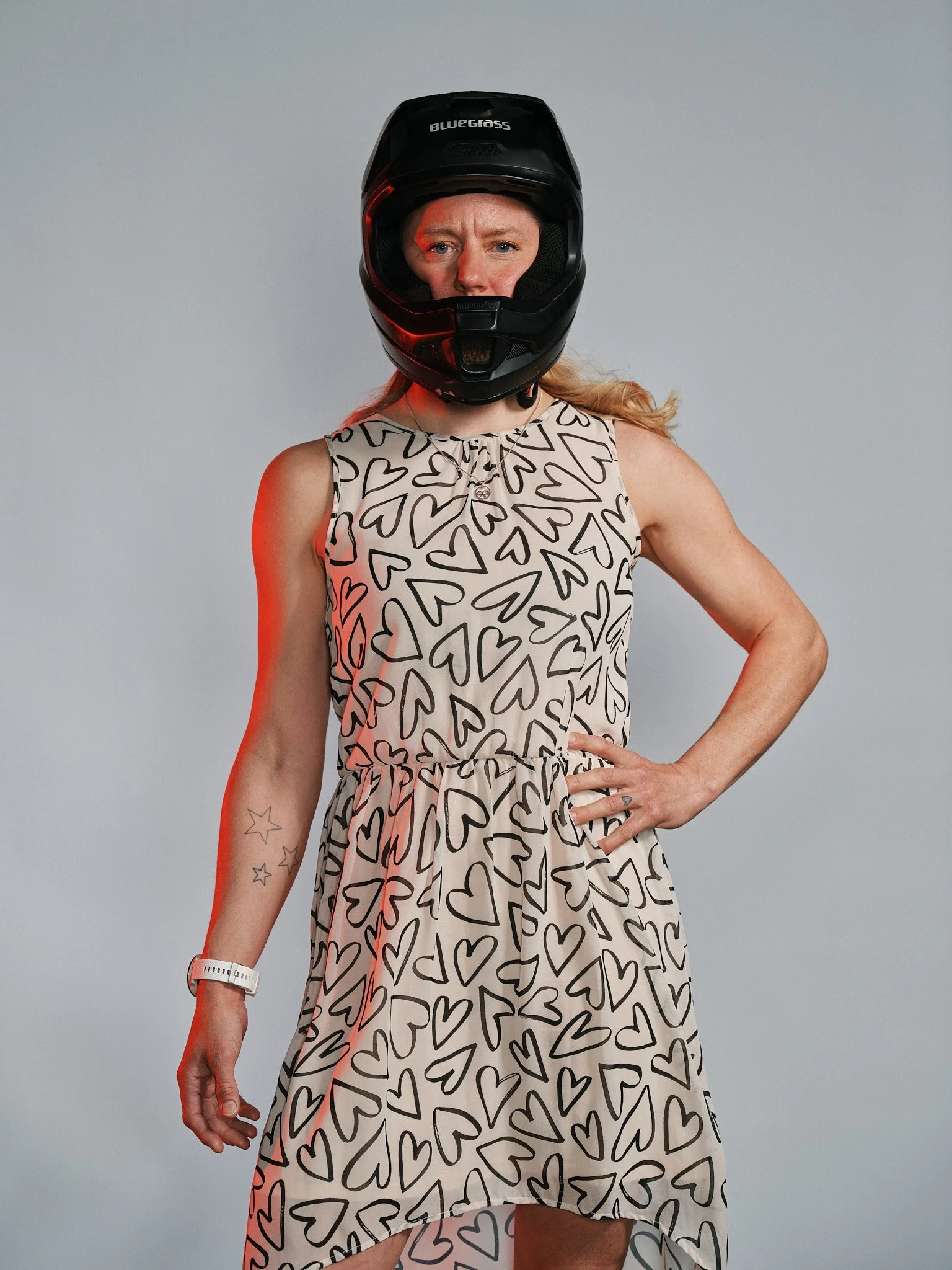 Donna con abito a fantasia di cuori, indossa un casco da moto nero, si trova davanti a uno sfondo grigio chiaro, con un polso watch bianco e tatuaggi di stelle sul braccio.