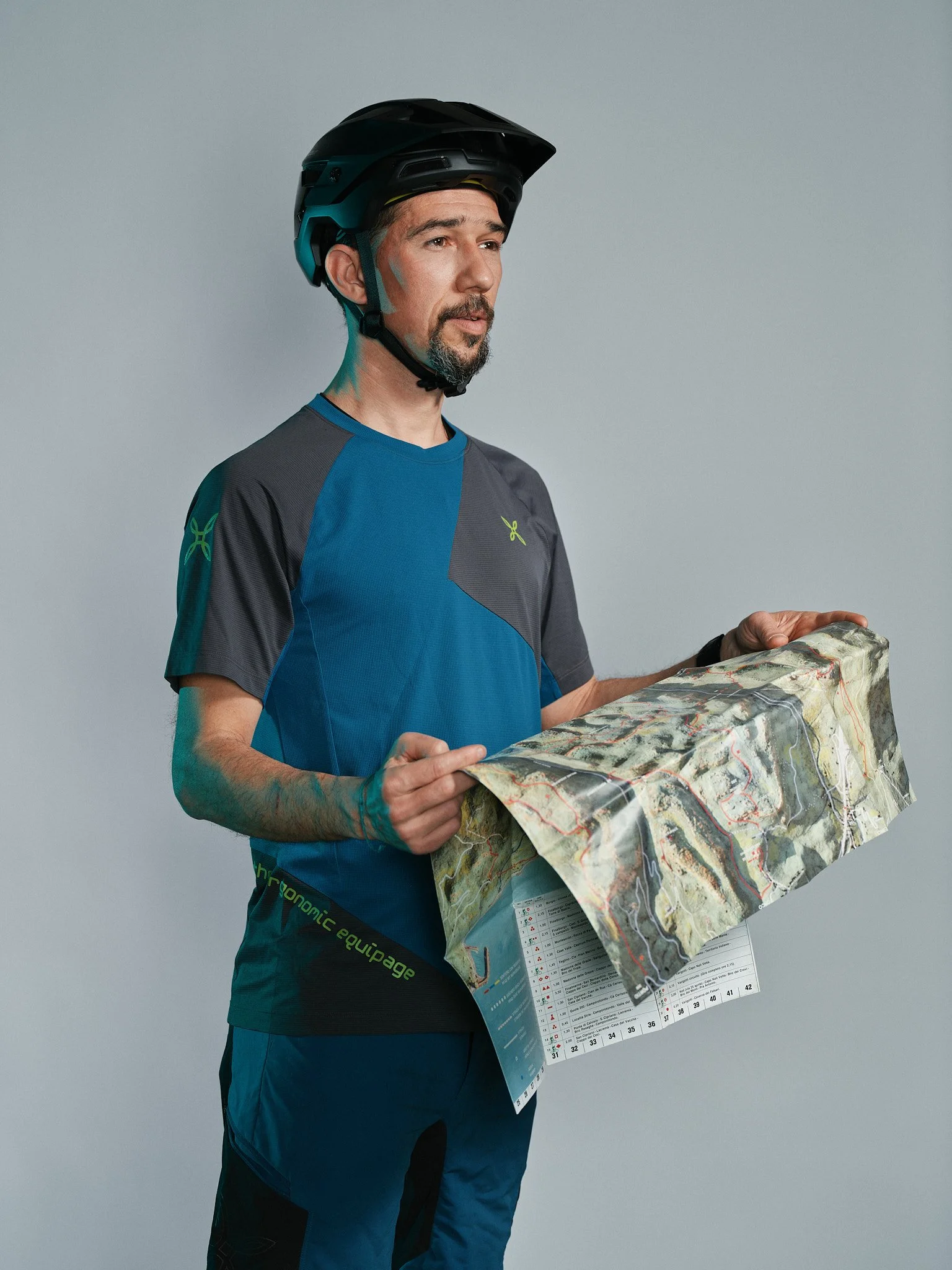 Uomo con caschetto da downhill, maglia tecnica e pantaloni da sport, che guarda una mappa topografica.