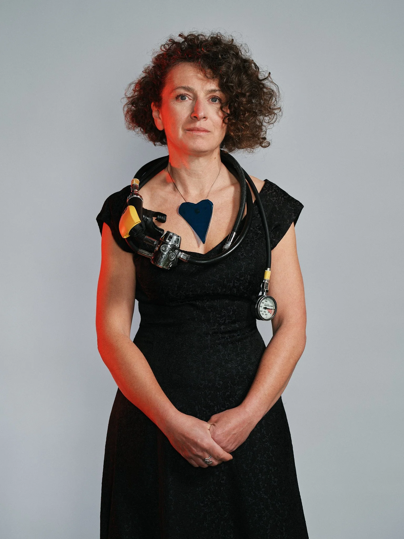 Una donna con capelli ricci, indossa una collana con un ciondolo a forma di cuore blu, una maschera di sub, un attrezzatura di subacquatica e un vestito nero. È in piedi contro uno sfondo grigio.