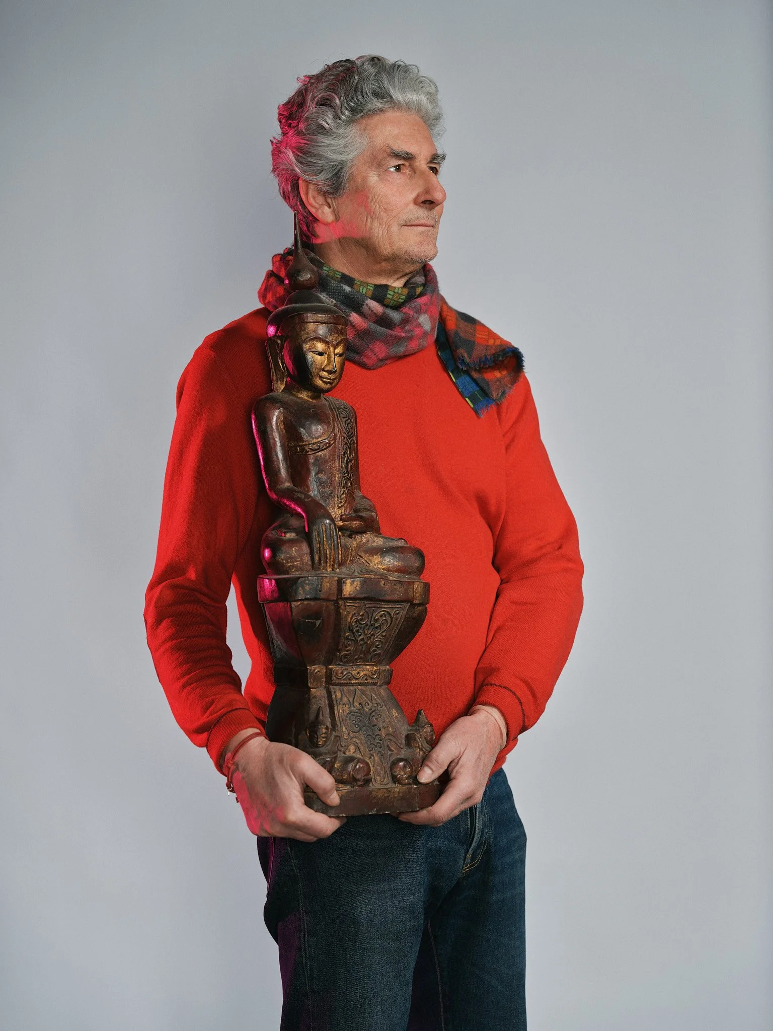 Uomo anziano che tiene una statua di legno di Buddha. Indossa una maglietta rossa e uno sciarpa a scacchi colorata. Sfondo grigio chiaro.