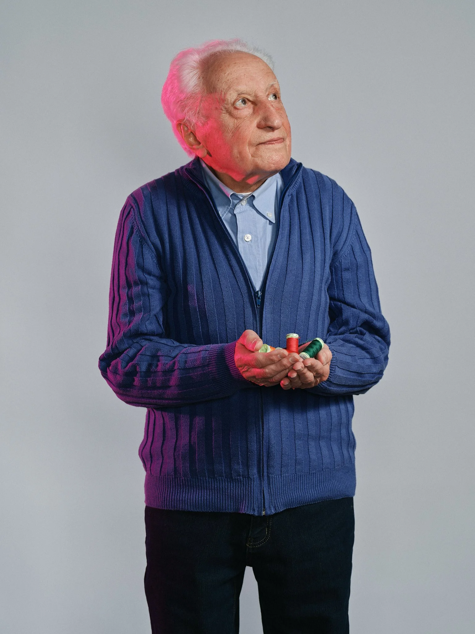 Uomo anziano con capelli bianchi, che indossa una maglia a zip blu e tiene spilli di diversi colori in mano, contro sfondo grigio.