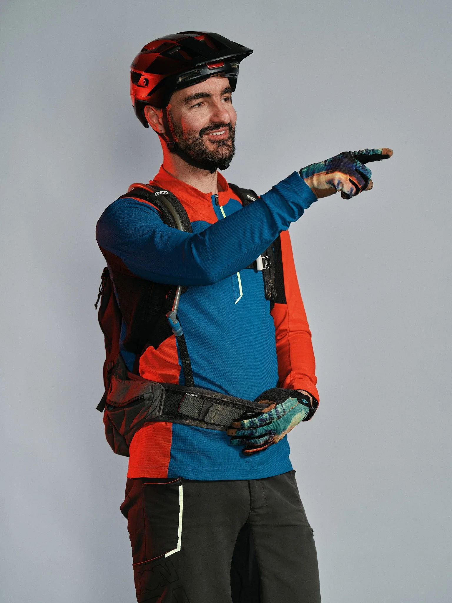 Uomo sorridente in abbigliamento da escursionismo, con casco, giacca sportiva, zaino, guanti e pantaloni, che indica o punta verso destra