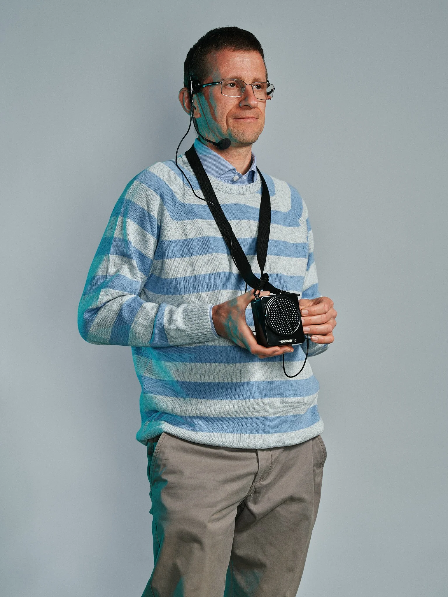 Uomo con occhiali e casco con microfono, indossa un maglione a righe blu e beige e tiene in mano una fotocamera.