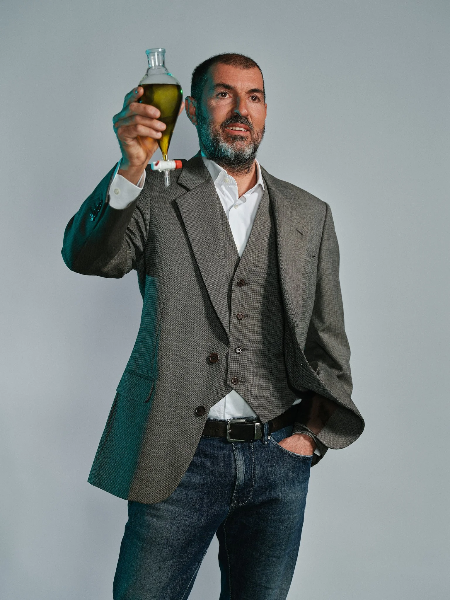 Un uomo con barba indossa una giacca grigia e jeans blu, tiene in mano un flask di vetro con un liquido verde, e ha una mano in tasca.