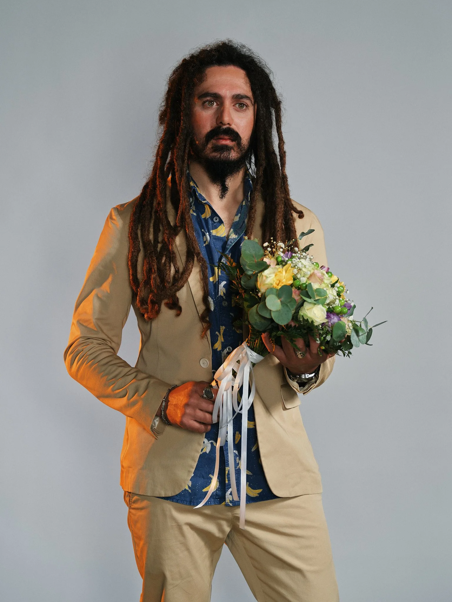 Uomo con dreadlocks che tiene un mazzo di fiori colorati e guarda verso l'obiettivo, indossa un abito beige e una maglietta con motivo di banana.