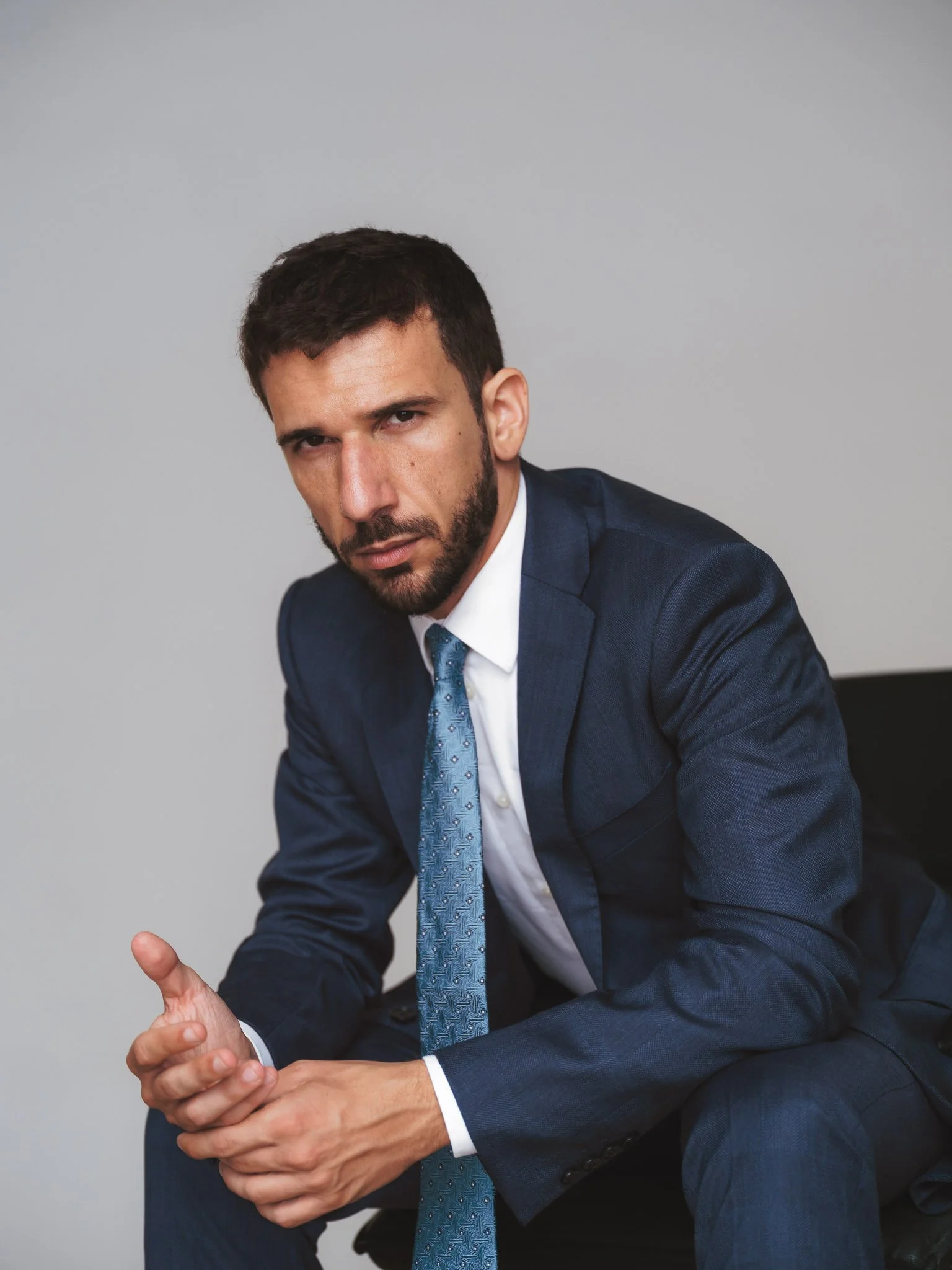 Uomo d'affari in giacca blu, cravatta azzurra, seduto con le mani in posizione di discussione o presentazione, sfondo grigio.