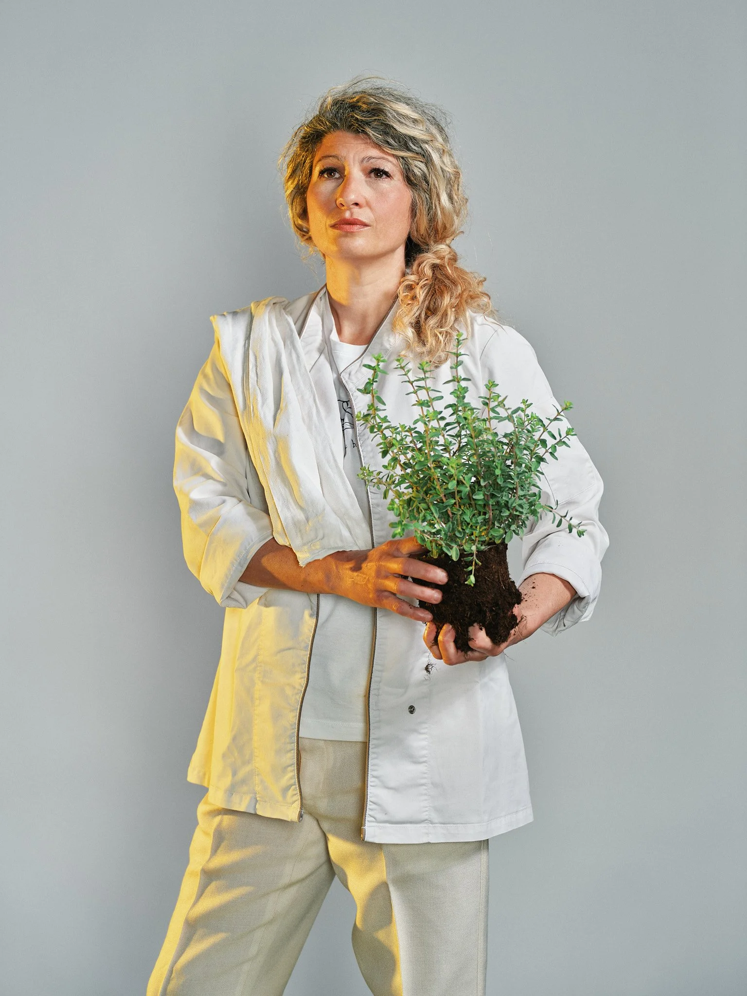 Una donna con capelli ricci e corti tiene un vaso con una pianta verde, indossa una giacca bianca e pantaloni beige, sfondo grigio.