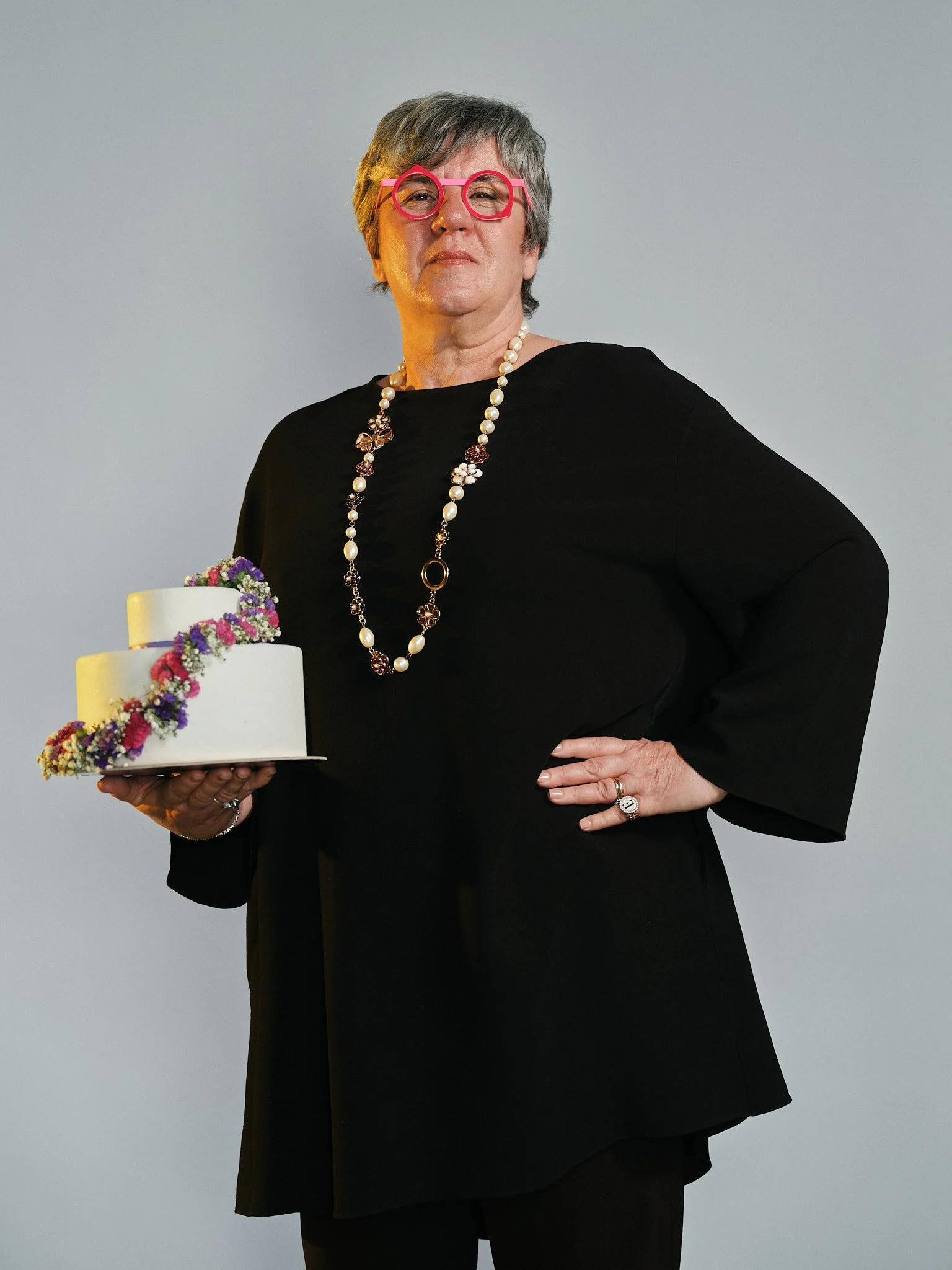 Una donna anziana con capelli grigi, indossa occhiali rossi, un vestito nero e una collana di perle. Tiene in mano una torta decorata con fiori colorati.