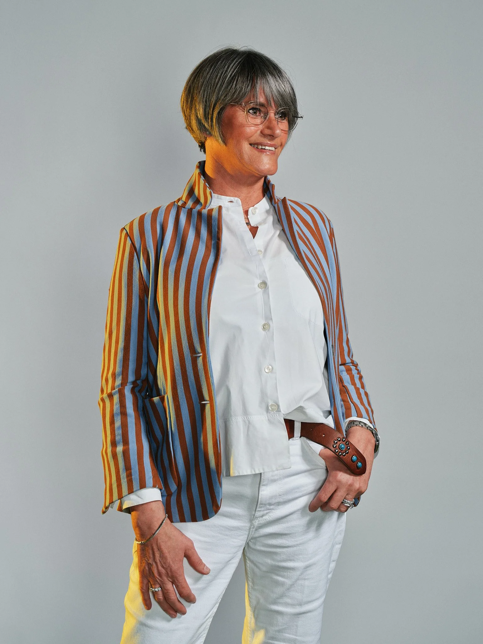 Donna con cappotto a righe colorate, camicia bianca, pantaloni bianchi e accessori colorati, posa con una mano sulla coscia.