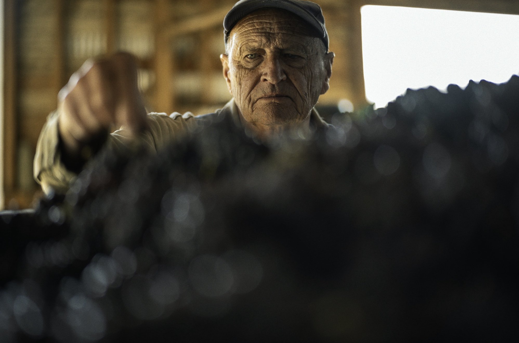 An elderly man with a serious expression, wearing a cap, is focused on working on something in front of him in a woodworking or crafting workshop.