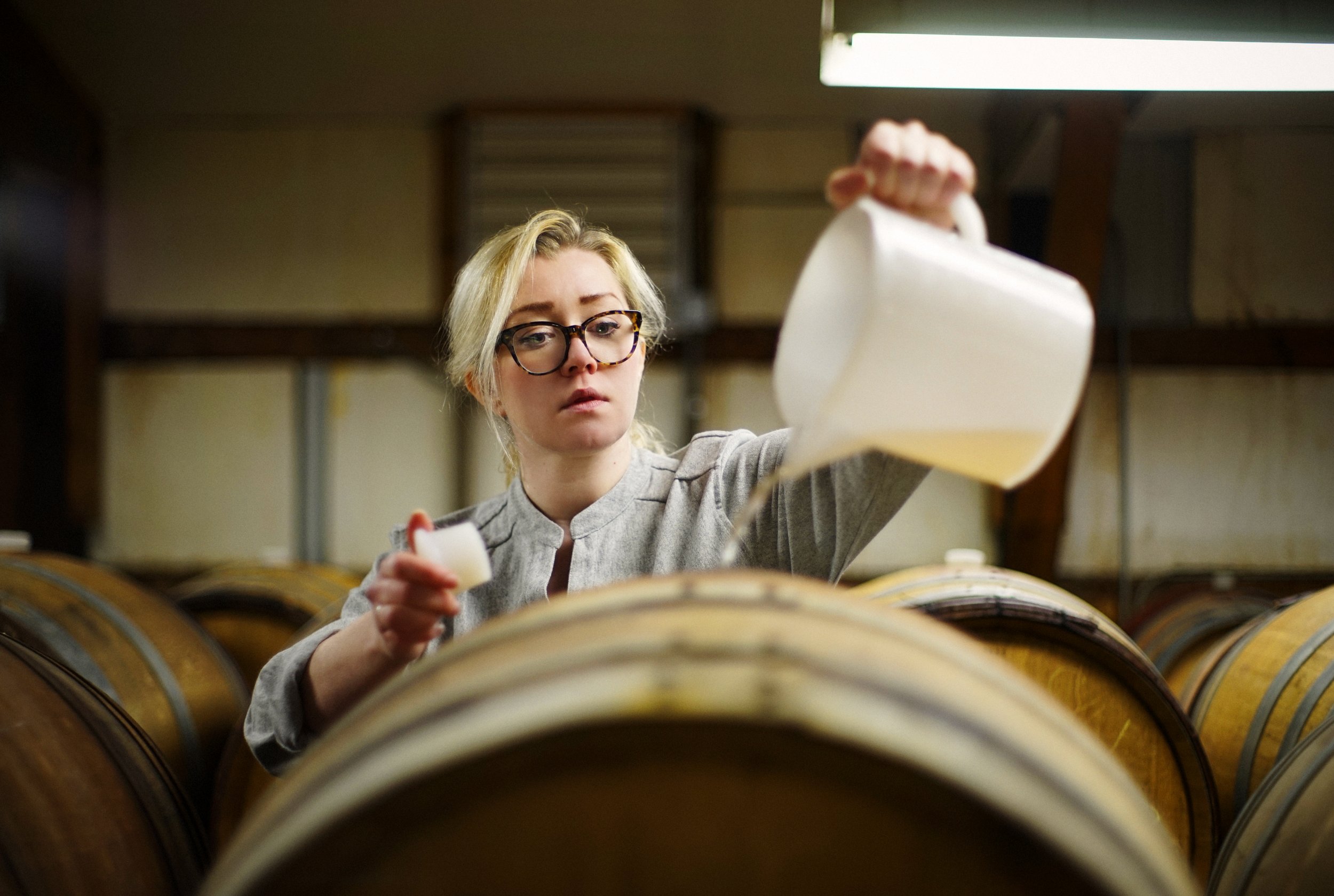 A woman with blonde hair and glasses pouring liquid from a white container into a large barrel in a cellar or winery with wooden barrels in the background.