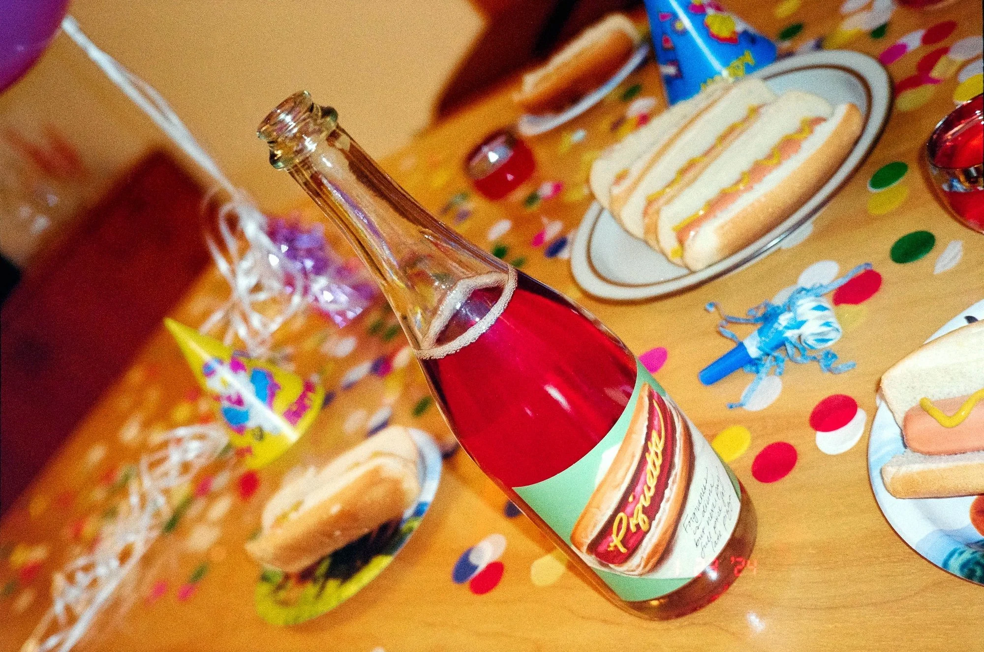 A party table with a bottle of Piquette, sandwich snacks, colorful confetti, scattered party hats, and birthday decorations.