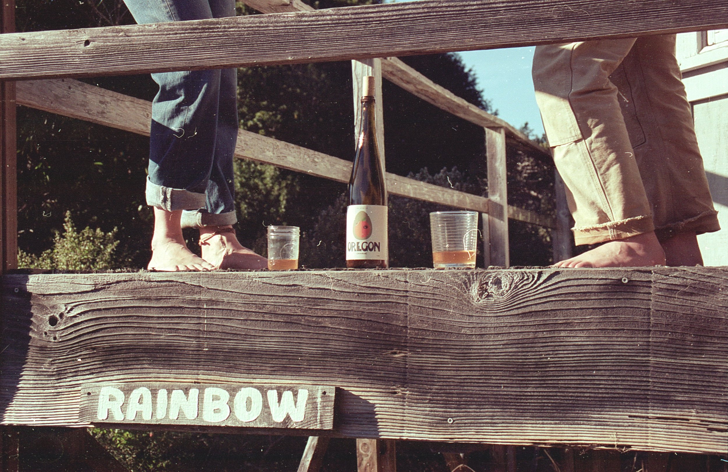 Two people standing on a wooden deck, one with rolled-up jeans and the other with rolled-up pants, with a bottle of our Oregon Orange wine.