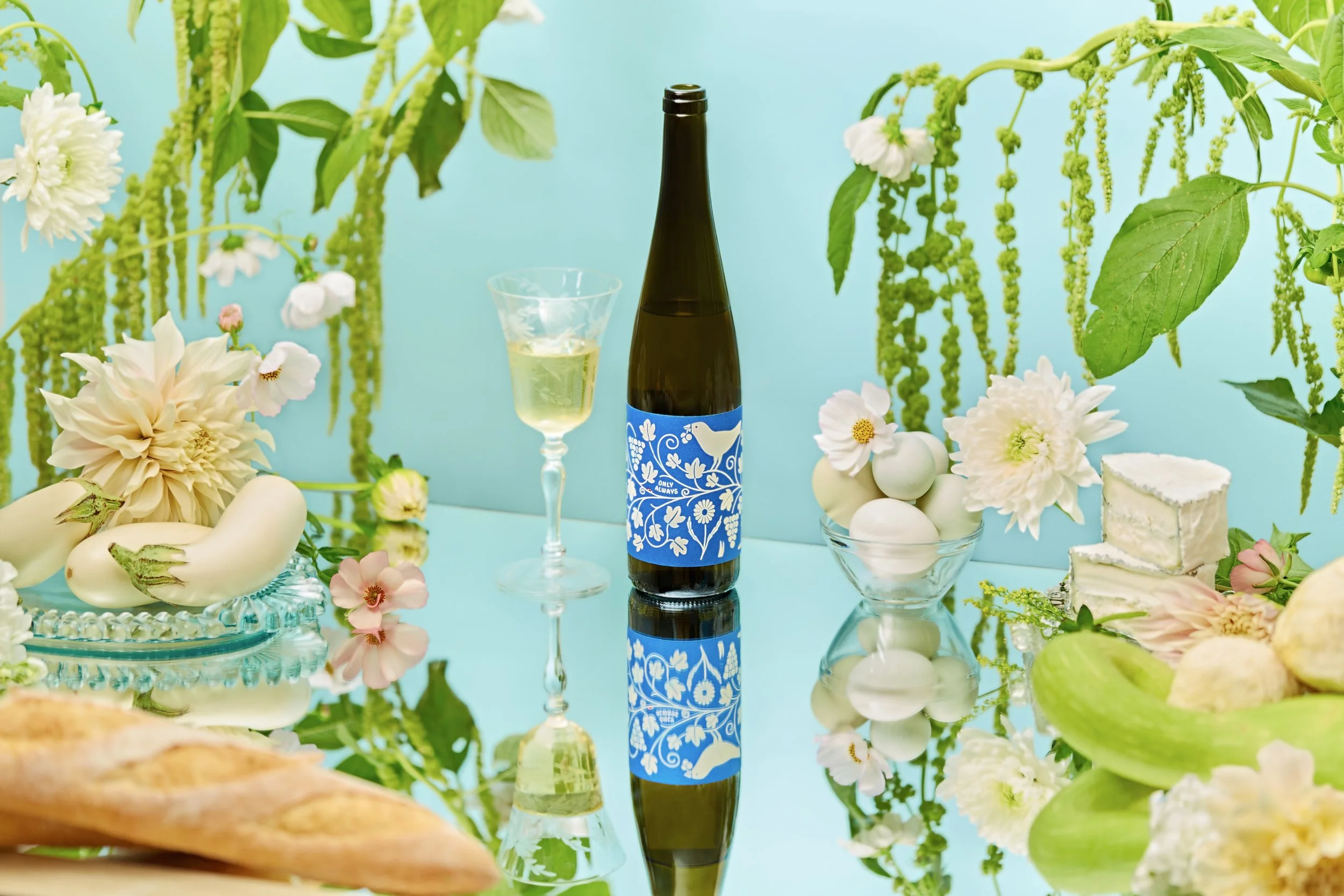 A still life setup with a bottle of wine, a glass of white wine, and various flowers and vegetables, including white eggplants and sliced bread, arranged on a reflective surface with hanging green plants and a light blue background.