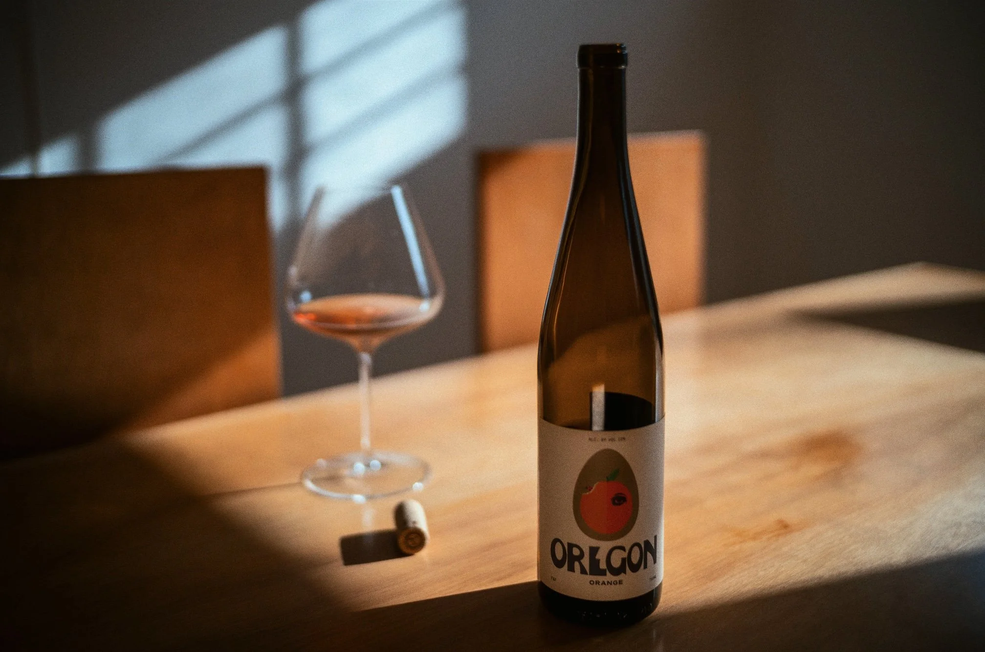A bottle of Oregon orange wine and a partially filled wine glass on a wooden table, with sunlight casting shadows on the wall and a cork lying on the table.