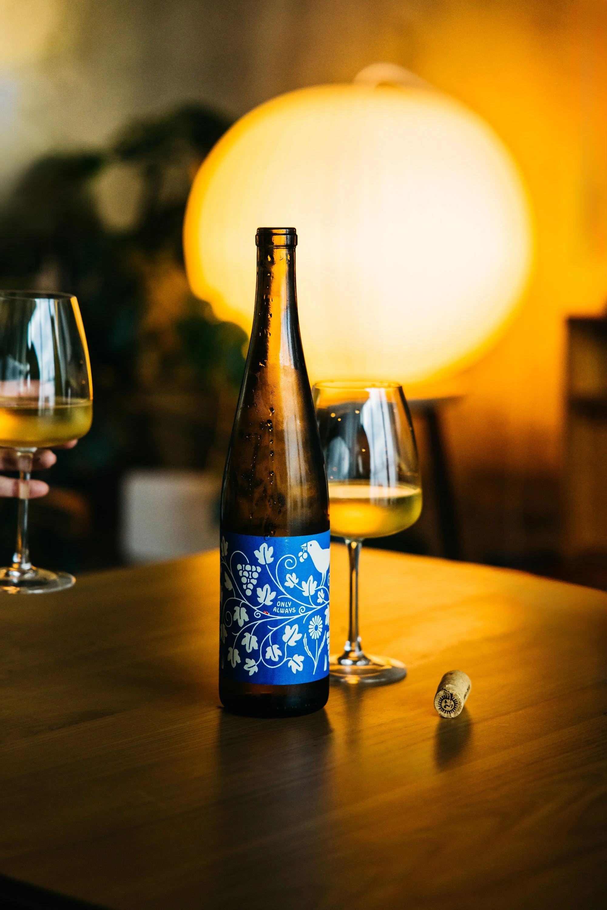 A bottle of our Only Always white wine, placed on a wooden table next to two wine glasses with white wine and a cork; a glowing round lamp in the background.