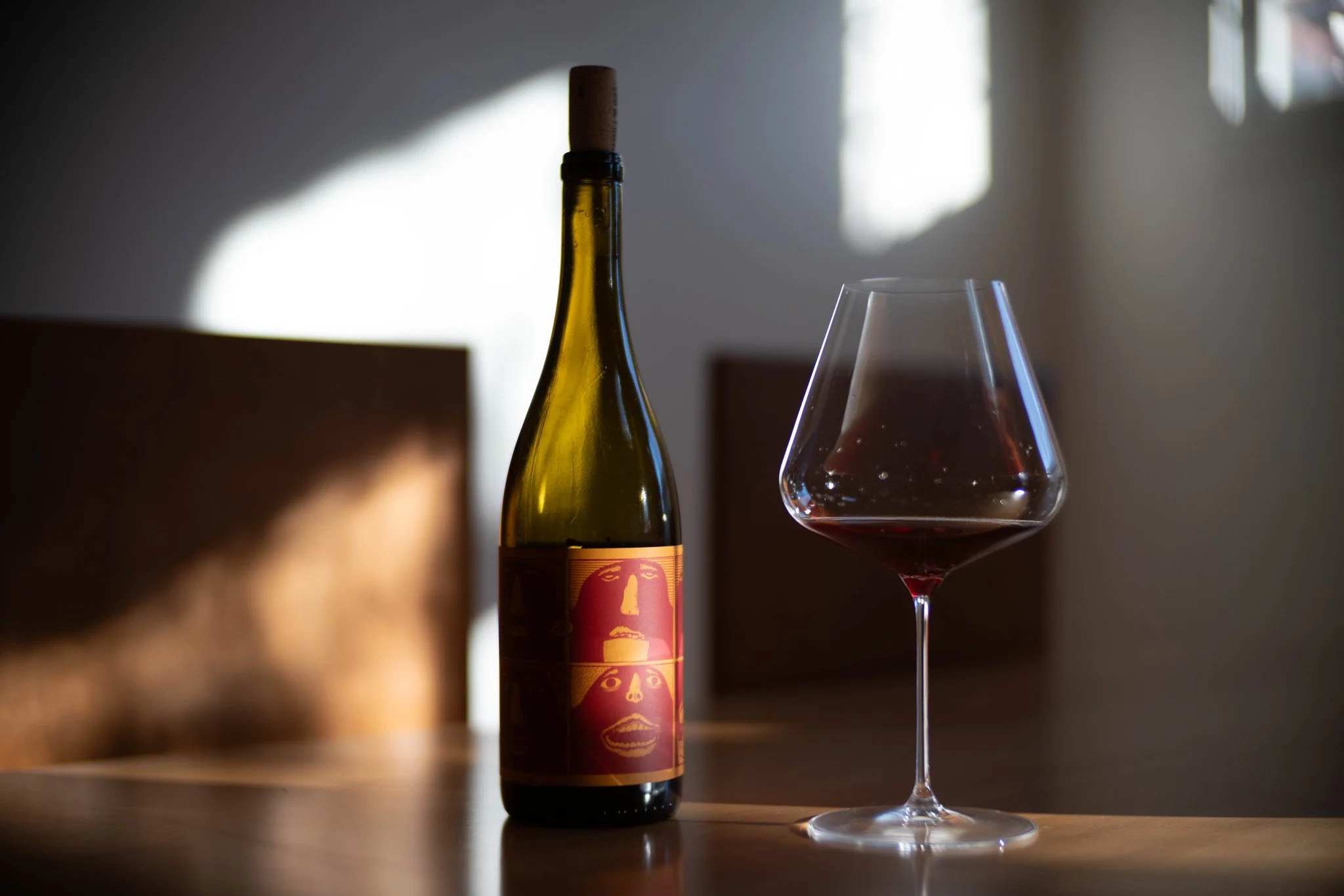 A bottle of our Willamette Valley Pinot Noir and a wine glass filled on a wooden table, with sunlight casting shadows in the background.