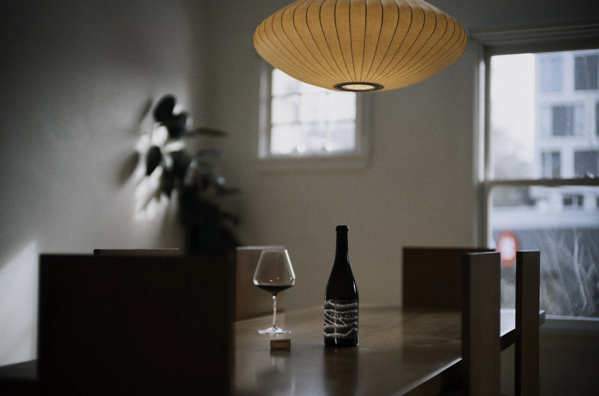 A dining table with a bottle of our Dark Wave red wine, a glass of red wine, and a cork. A paper lantern hanging from the ceiling. Sunlight coming through the windows in a modern apartment.
